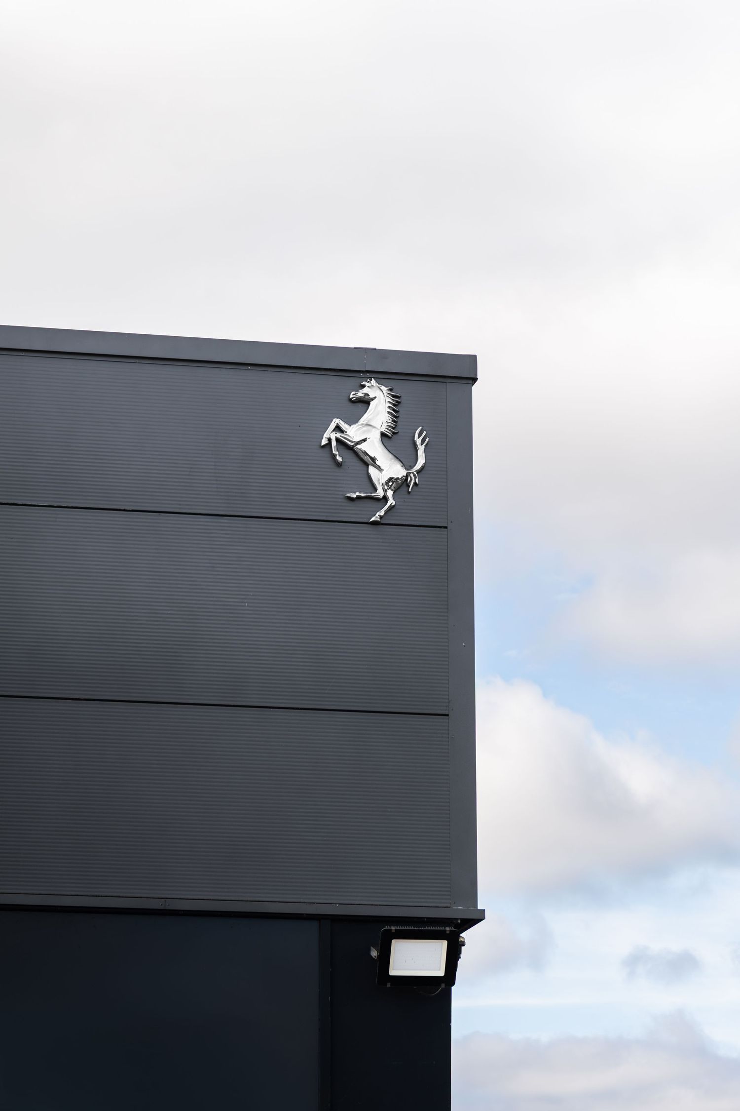 Ferrari prancing horse logo on dark gray building exterior against cloudy sky.