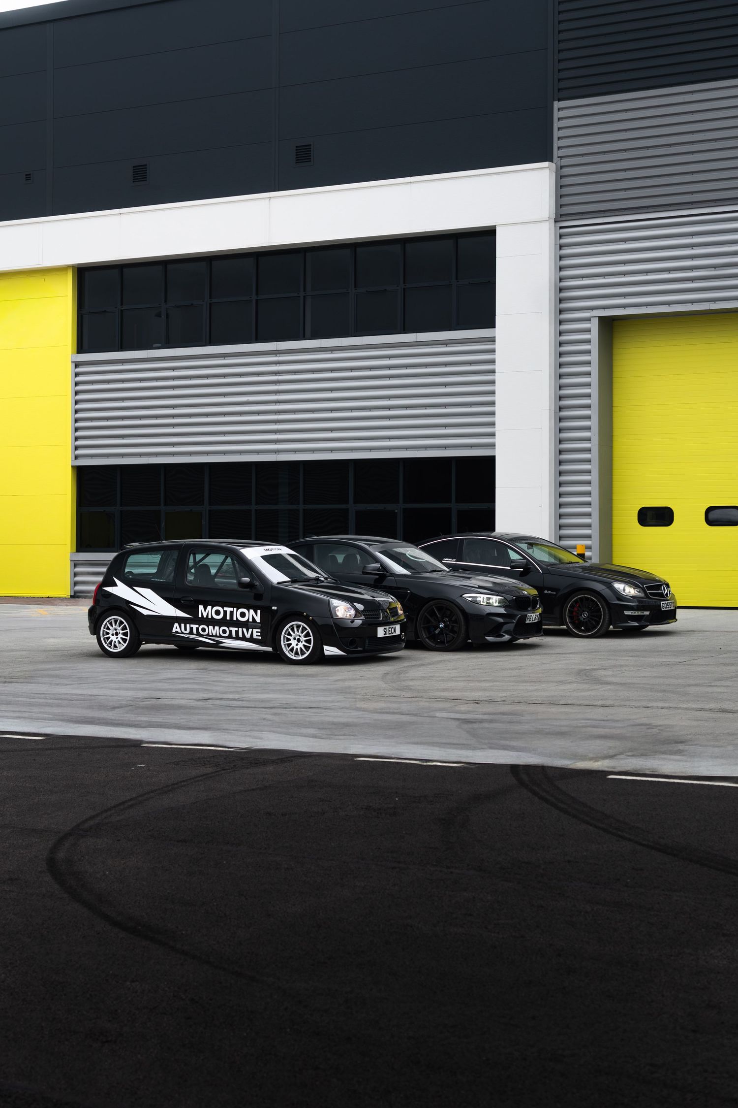 Three black Motion Automotive cars parked outside a modern industrial unit with yellow roller shutter doors.