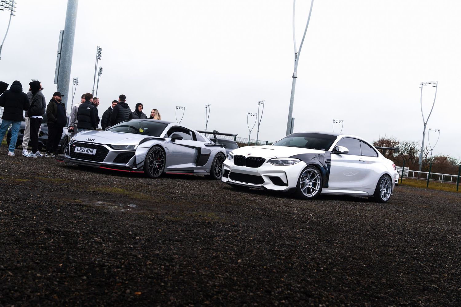White Audi R8 GT and white BMW M2 parked side by side at an outdoor car show.
