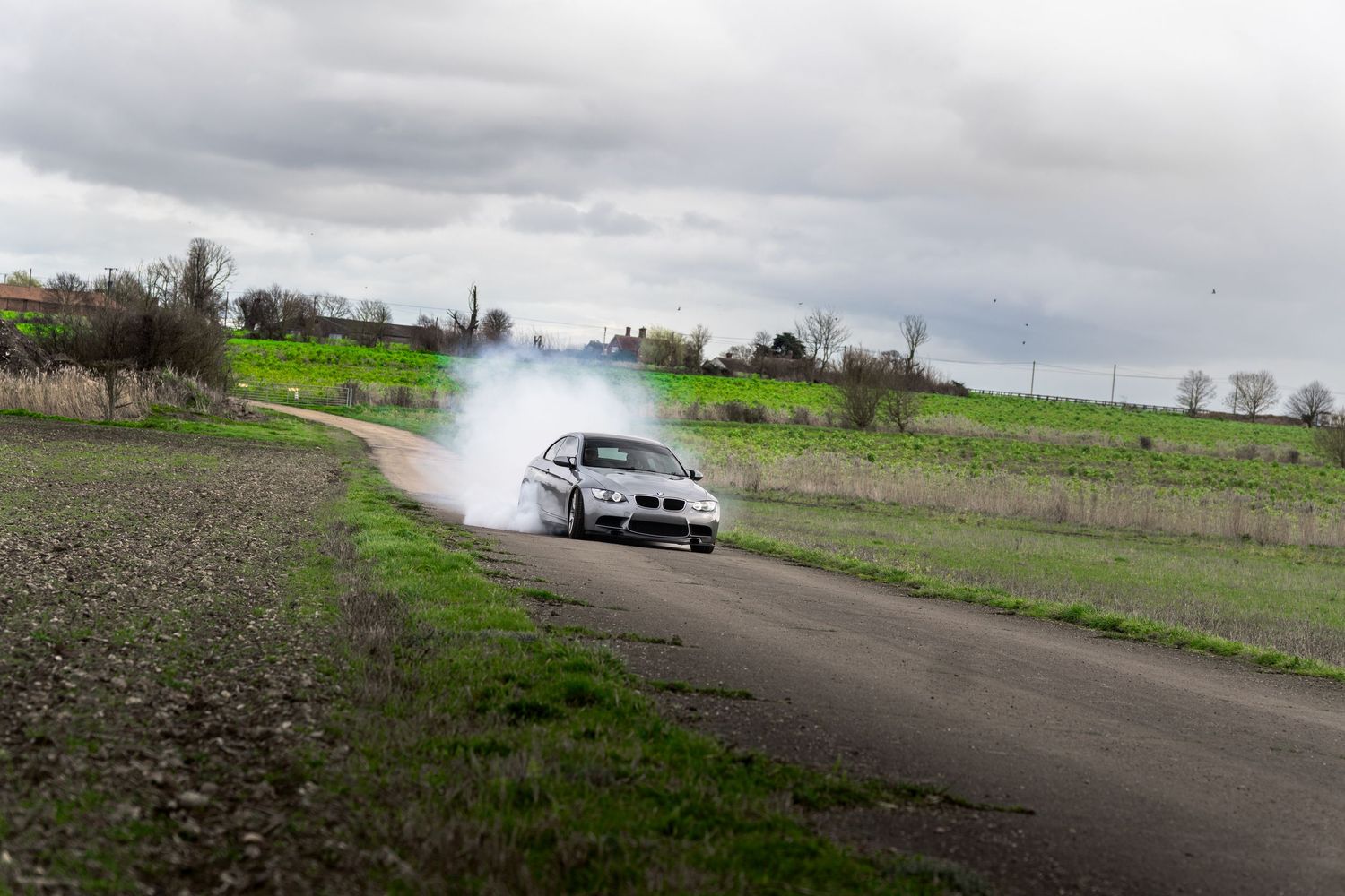 Silver BMW M3 coupe performing a dramatic drift with tire smoke on a rural countryside road.