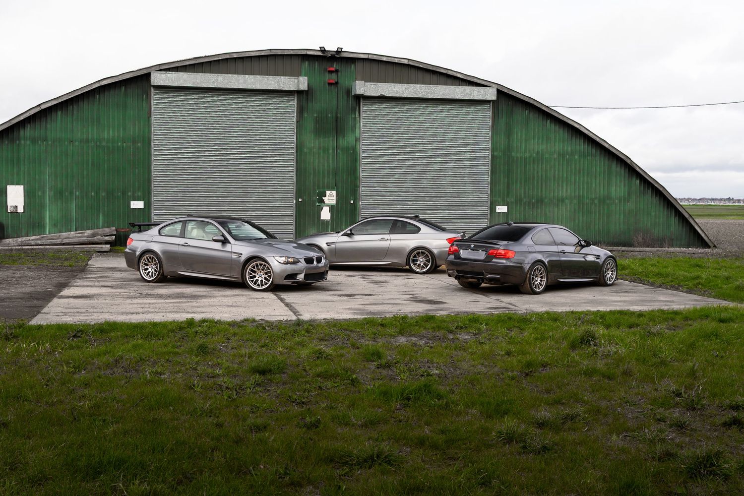 Three silver BMW M3 coupes parked in front of a large green industrial hangar building.
