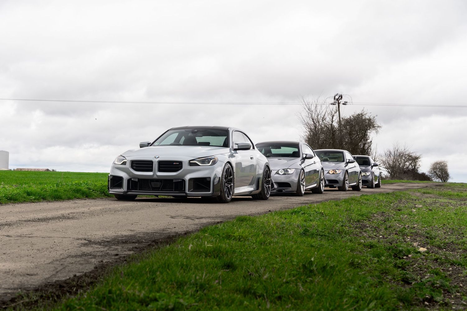 Three silver BMW M2 and M3 coupes lined up on a countryside road under overcast skies.
