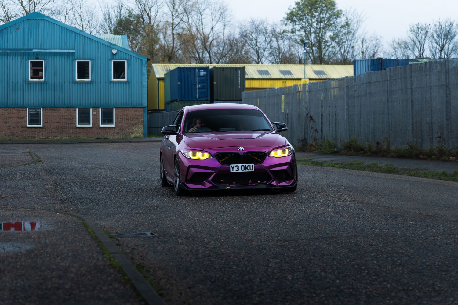 Purple BMW M2 with yellow headlights parked in an industrial area.