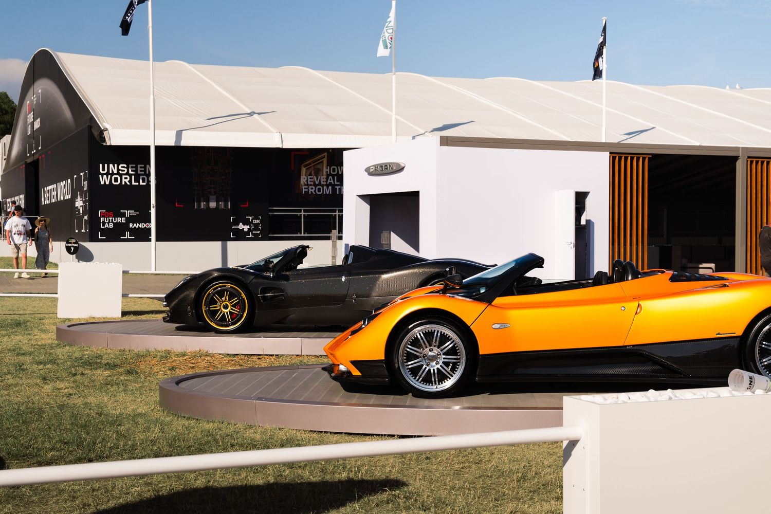 Two luxury supercars, black and orange convertibles, displayed outside a motorsport exhibition tent at an outdoor event.