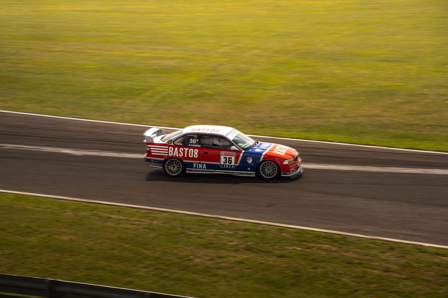 Bastos-sponsored BMW touring car racing on a motorsport circuit at sunset.