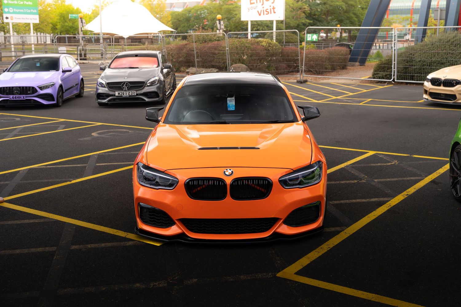Front view of a bright orange BMW 1 Series with custom wide body kit in a yellow-lined parking lot.