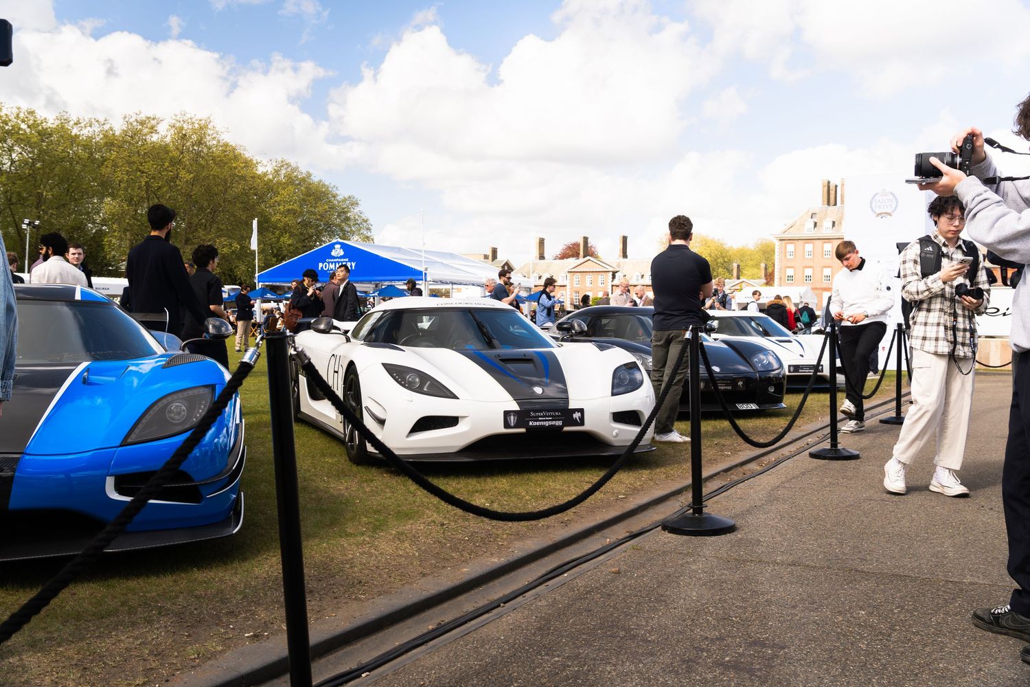 White Koenigsegg Agera and blue supercar displayed behind rope barriers at outdoor car show.