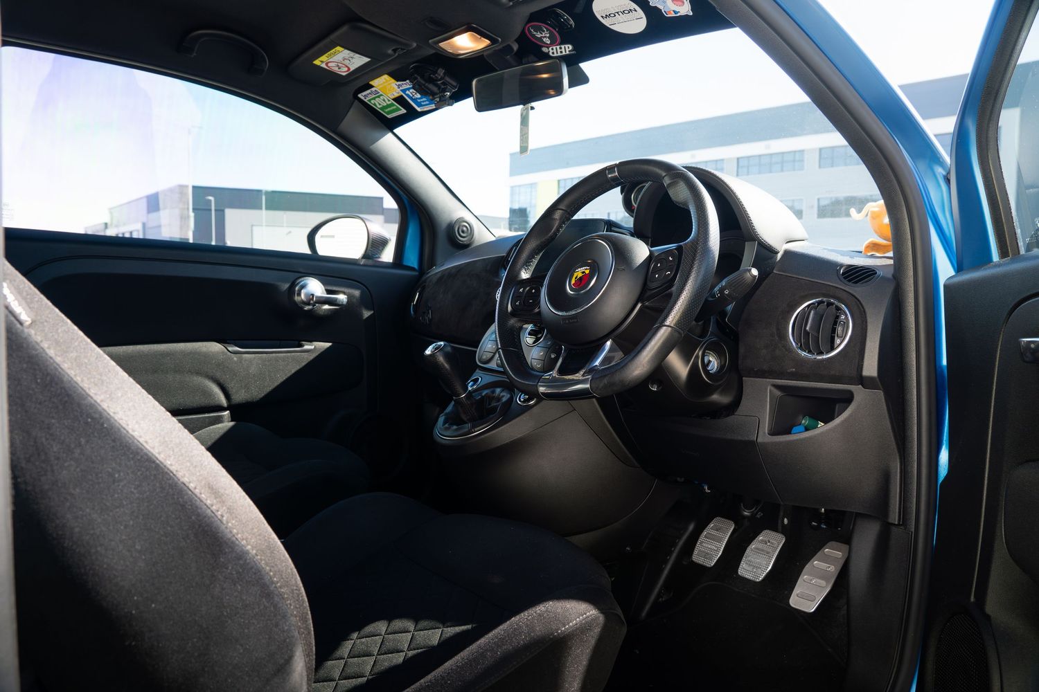 Interior of a blue Abarth 595 showing sport steering wheel, aluminum pedals, and black bucket seats.