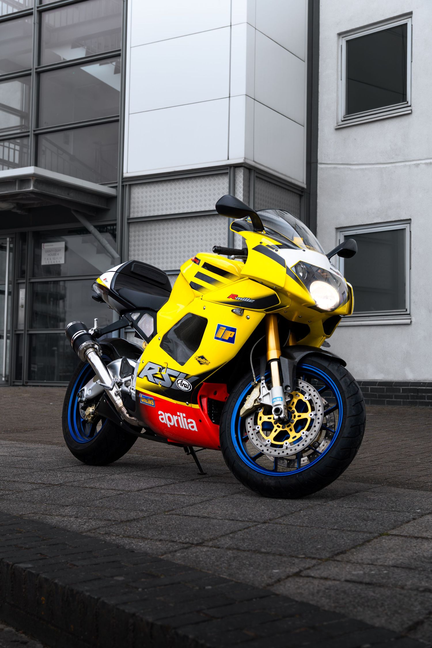 Yellow Aprilia RS5 sport motorcycle with blue wheels parked outside a modern building.