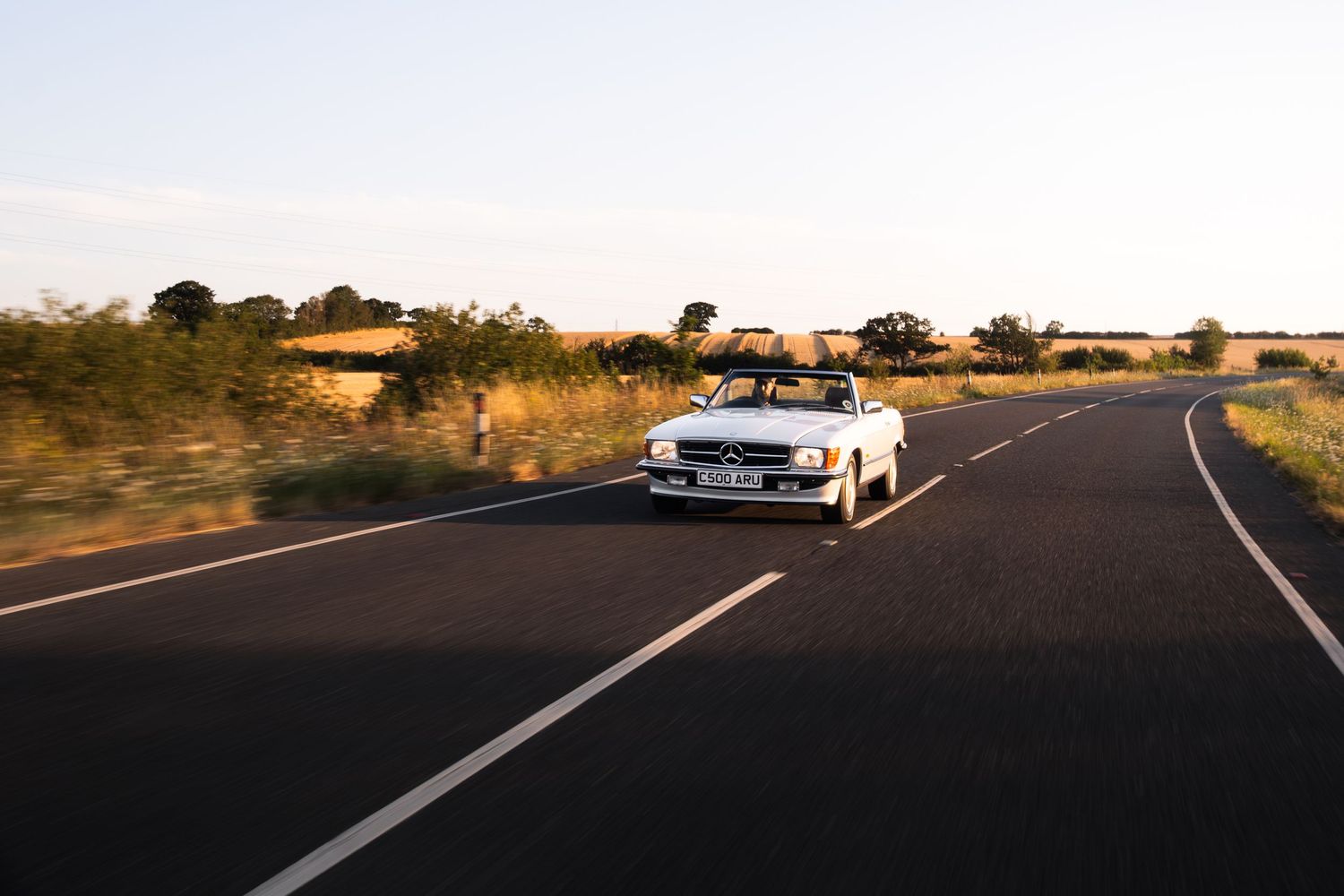 White convertible Mercedes driving on a scenic rural road at golden hour sunset.