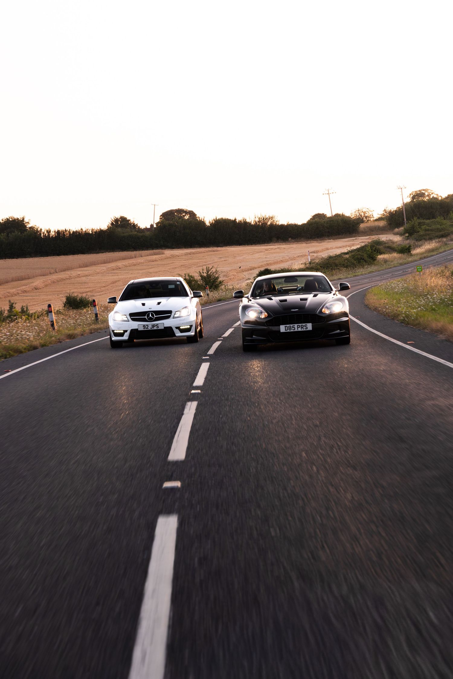 Two luxury sports cars, a white Mercedes and dark Aston Martin, racing side by side on a rural road at dusk.