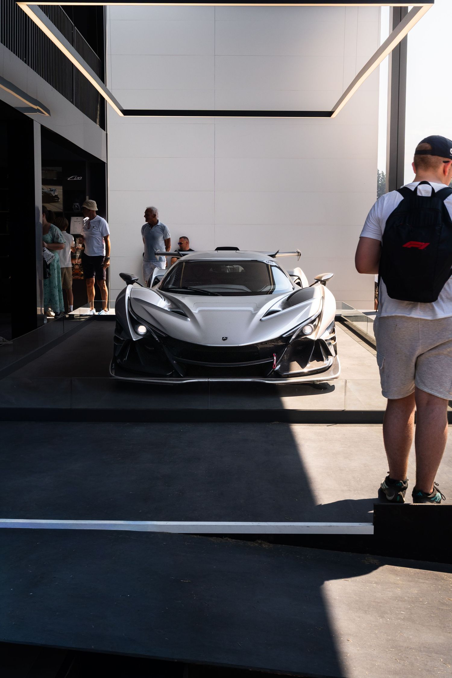White hypercar displayed in a pit garage at a racing event, surrounded by spectators admiring the exotic supercar.