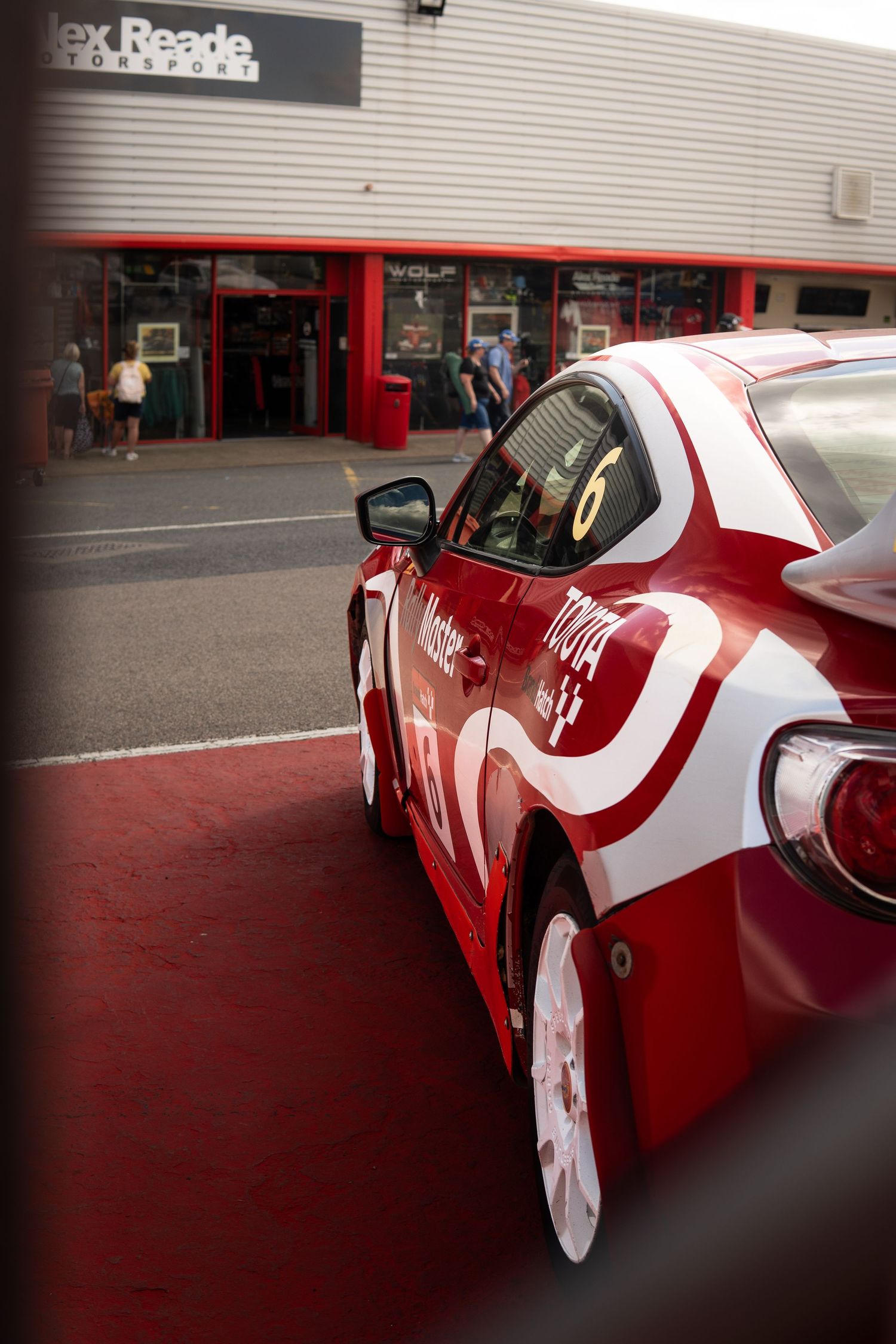 Red and white number 6 Toyota racing car parked outside Alex Reade Motorsport garage.