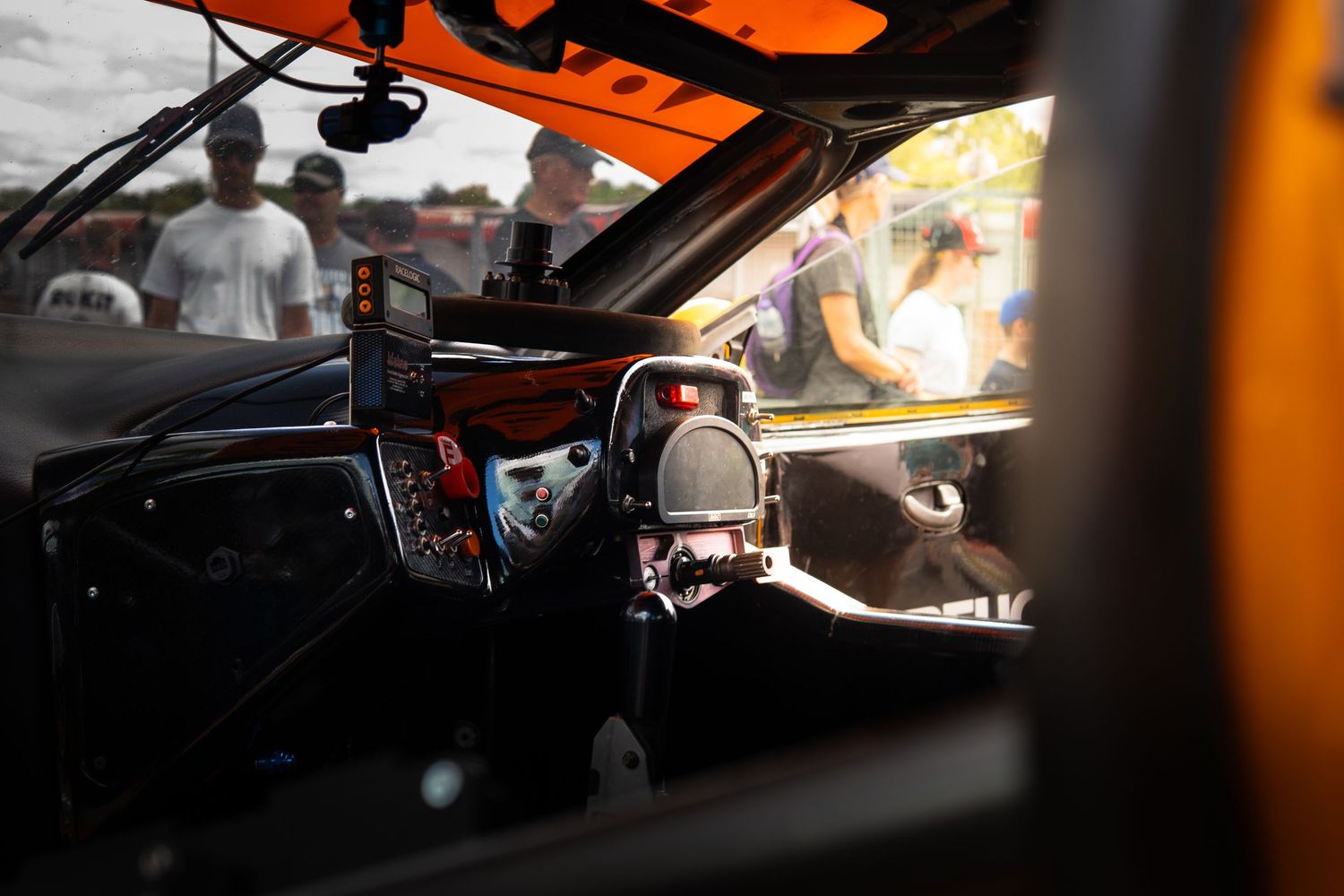 Interior view of an orange race car cockpit showing steering wheel and dashboard with spectators outside.