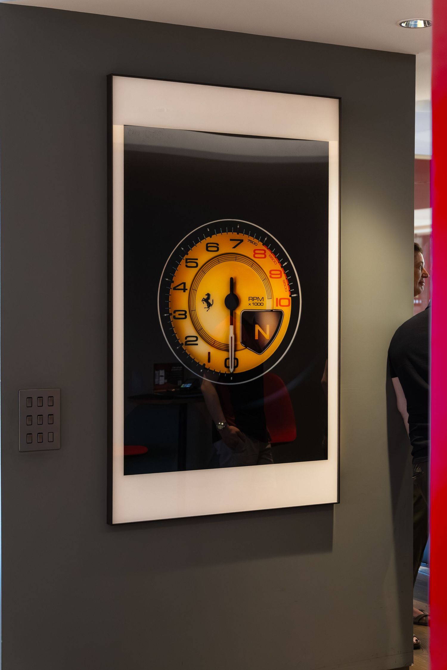 Ferrari tachometer gauge art print displayed on a dark gray wall in a modern showroom setting.