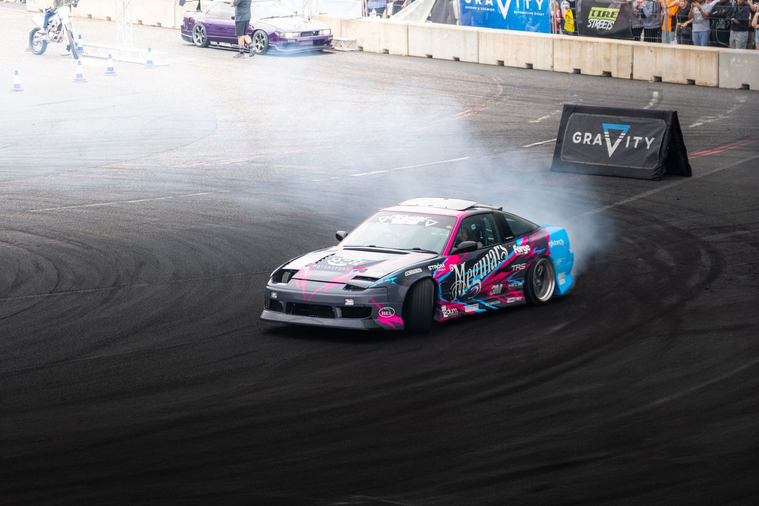 A pink and black drift car produces heavy tire smoke while drifting at a Gravity motorsport event at night.