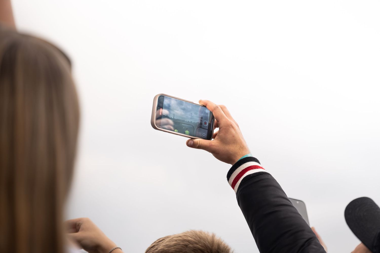 Person holding a smartphone above a crowd to photograph an event against an overcast sky.