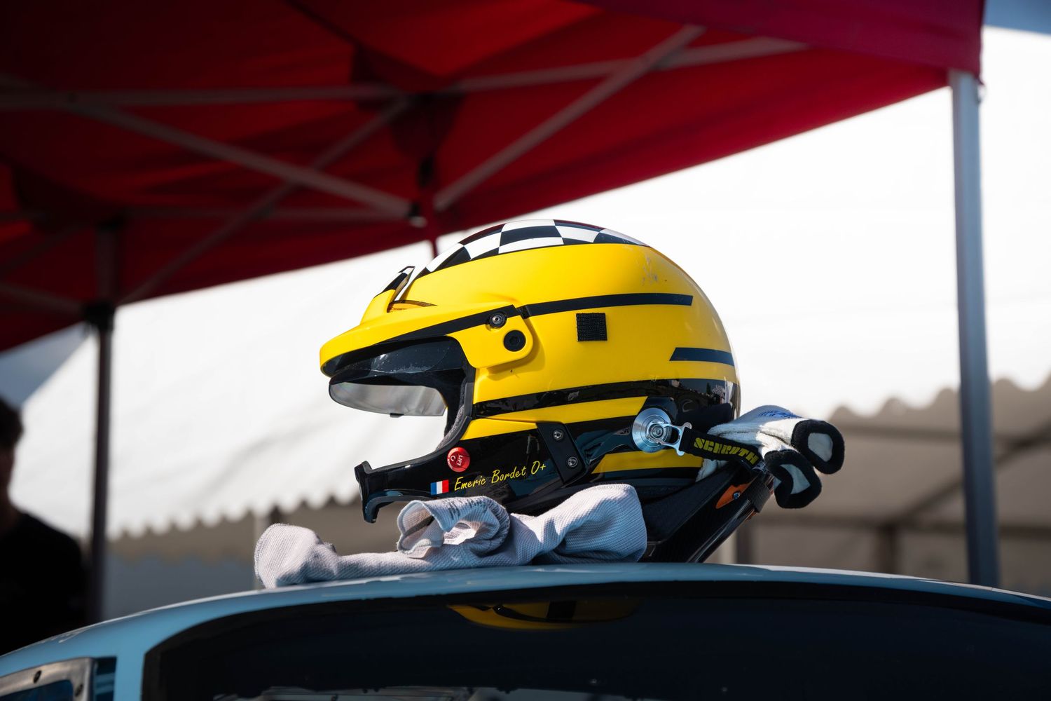 Yellow racing helmet resting on car roof under red canopy at motorsport event.