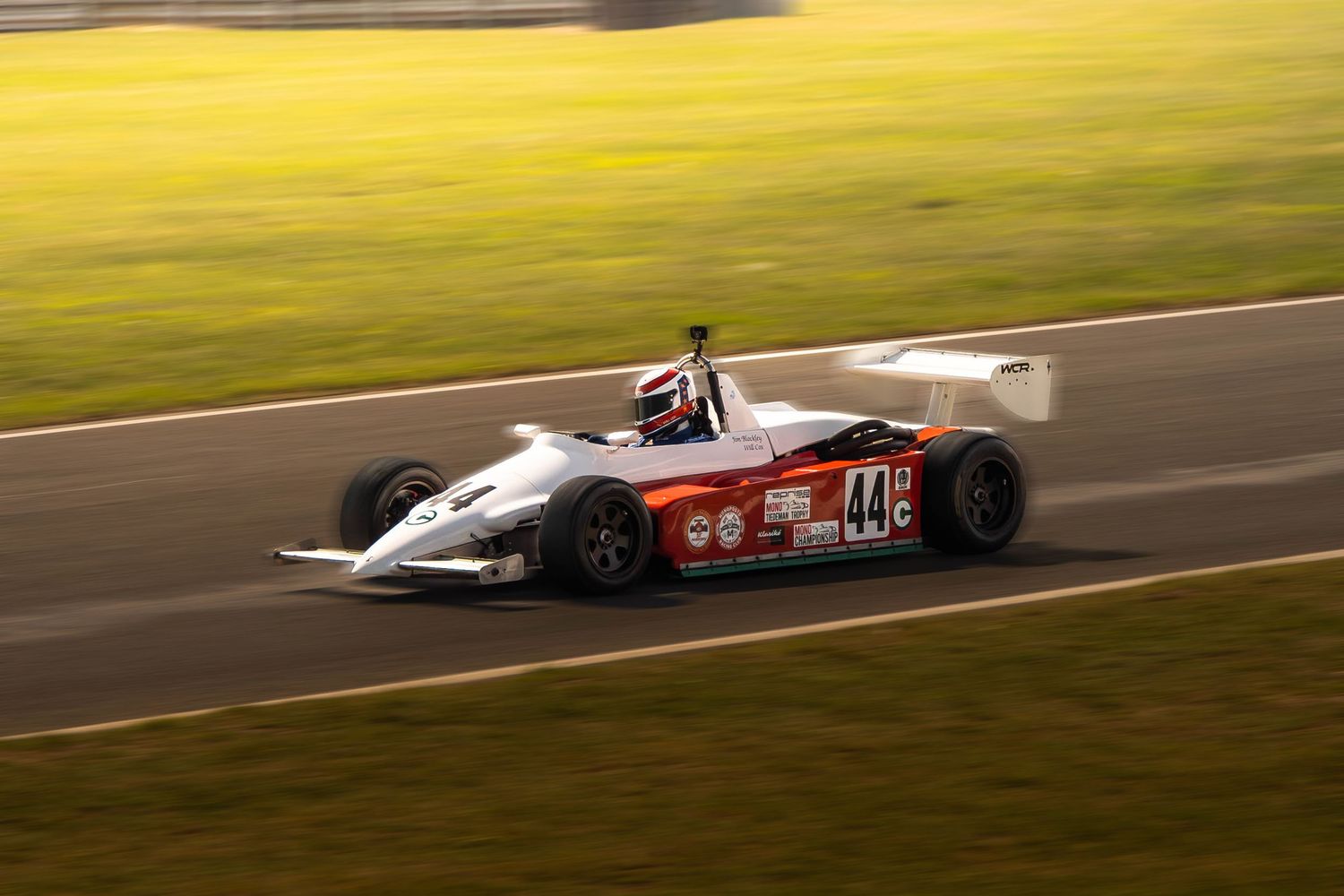 White and red Formula racing car number 44 speeds along a sunlit motorsport circuit.