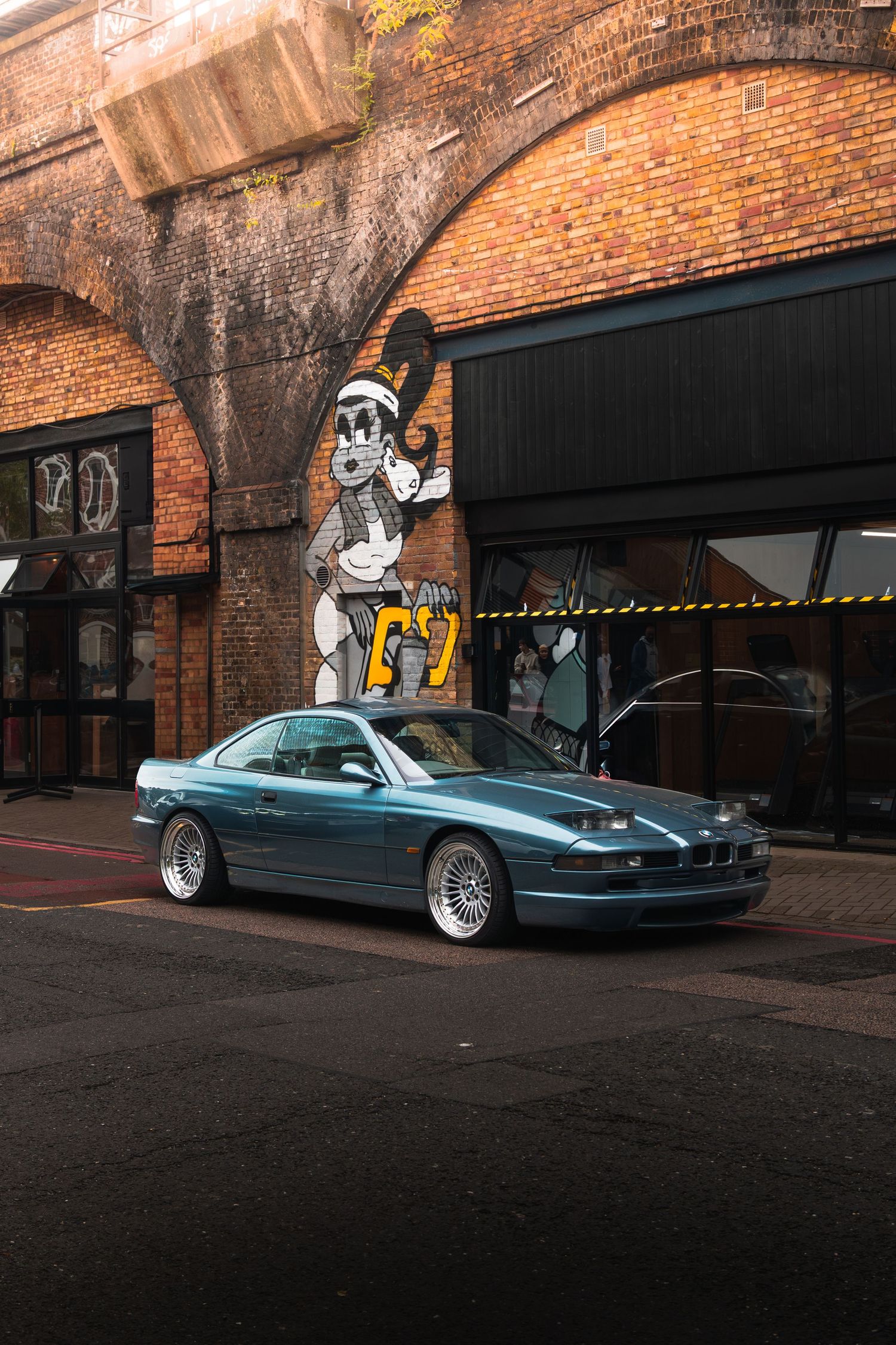 A teal BMW 8 Series coupe parked outside a brick building with cartoon graffiti mural.