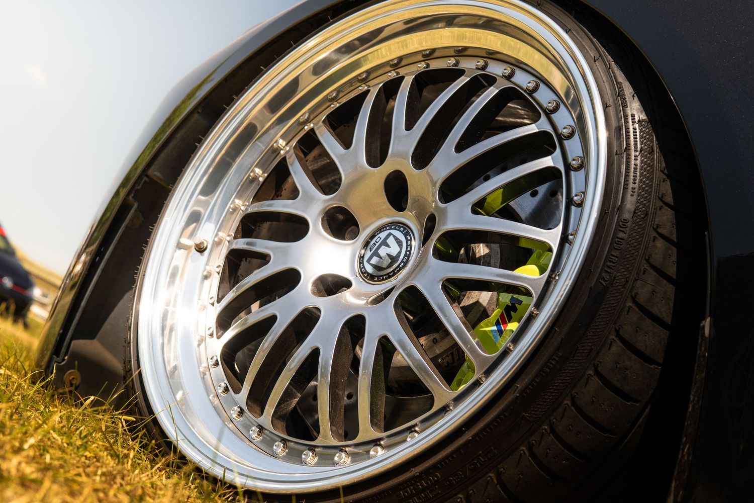 Close-up of polished three-piece mesh alloy wheel on lowered black car on grass.