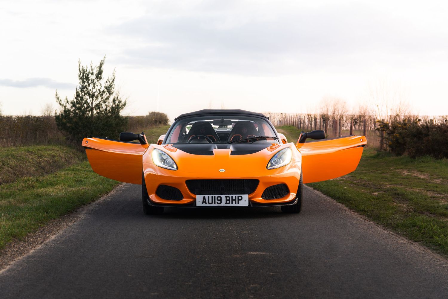Front view of orange Lotus Elise with doors open on a rural country road at dusk.