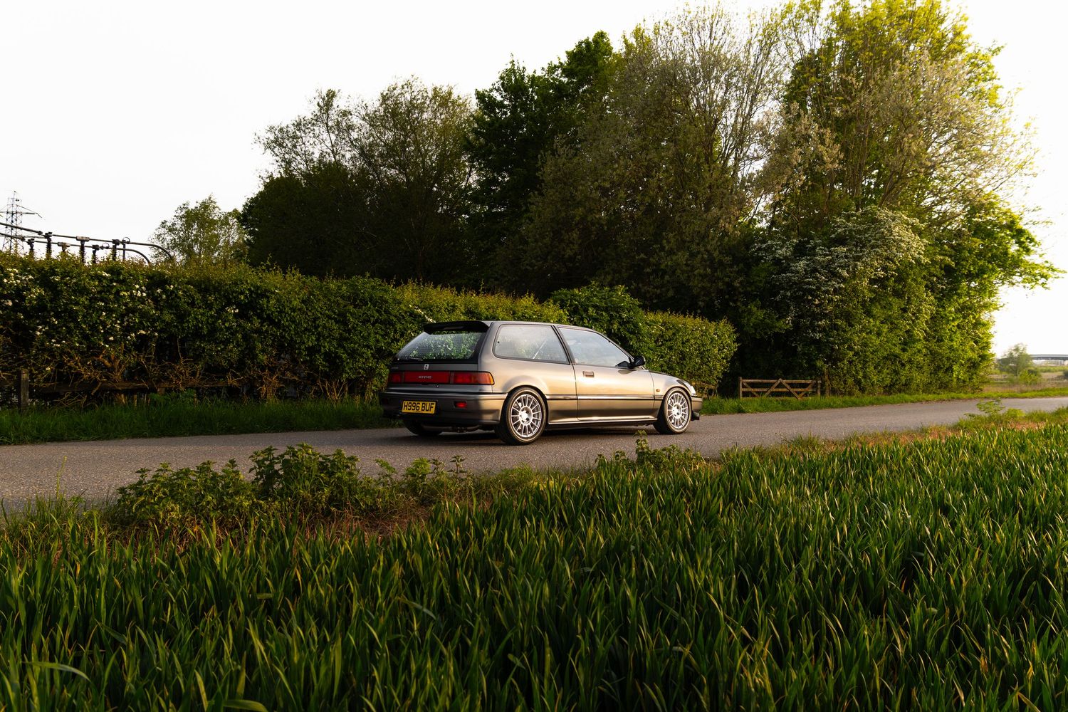 Rear three-quarter view of a lowered silver Honda Civic EG on a rural road with green hedges at golden hour.