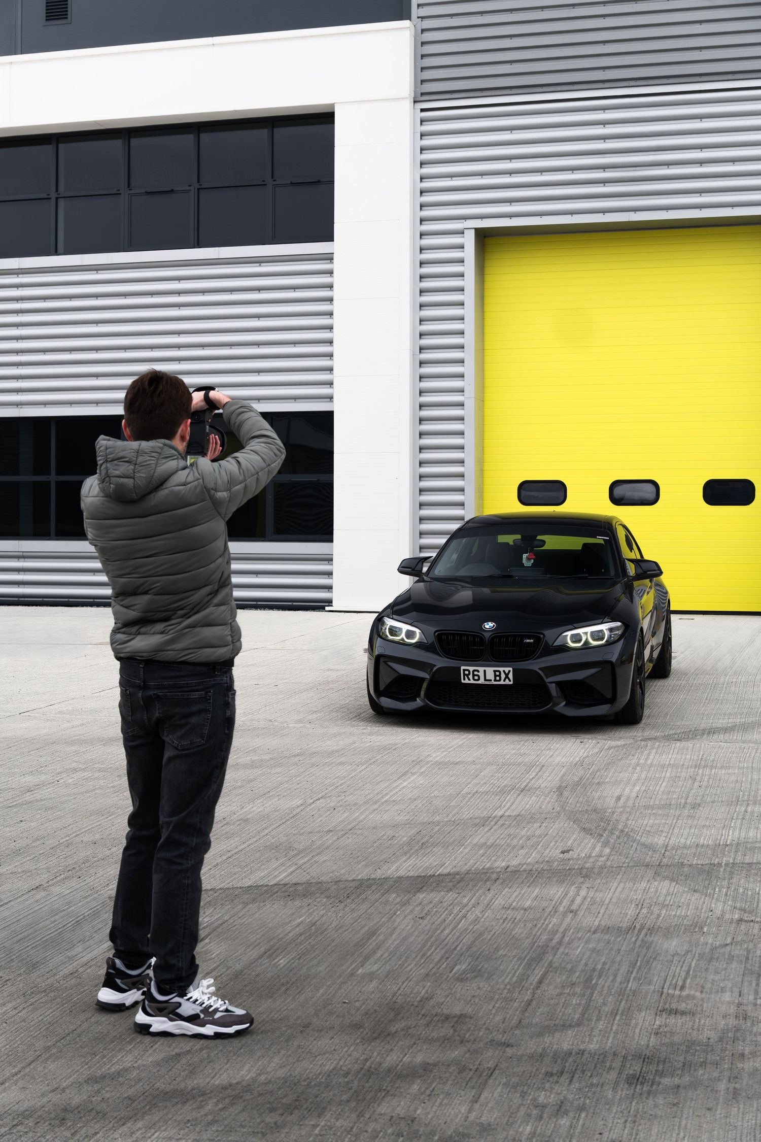 Photographer capturing a black BMW M2 coupe parked in front of yellow garage doors.