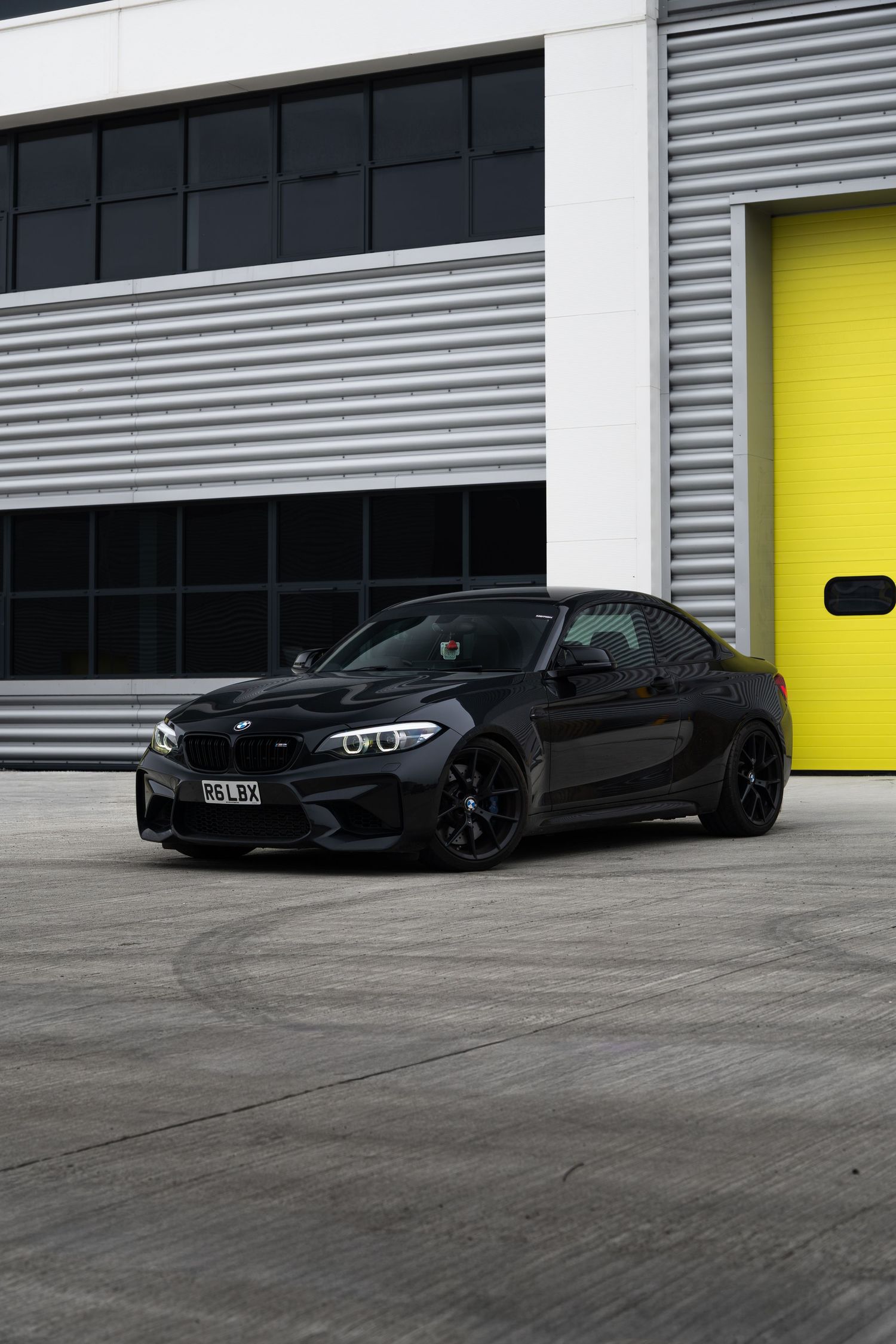 Black BMW M2 coupe parked outside an industrial building with yellow garage doors.