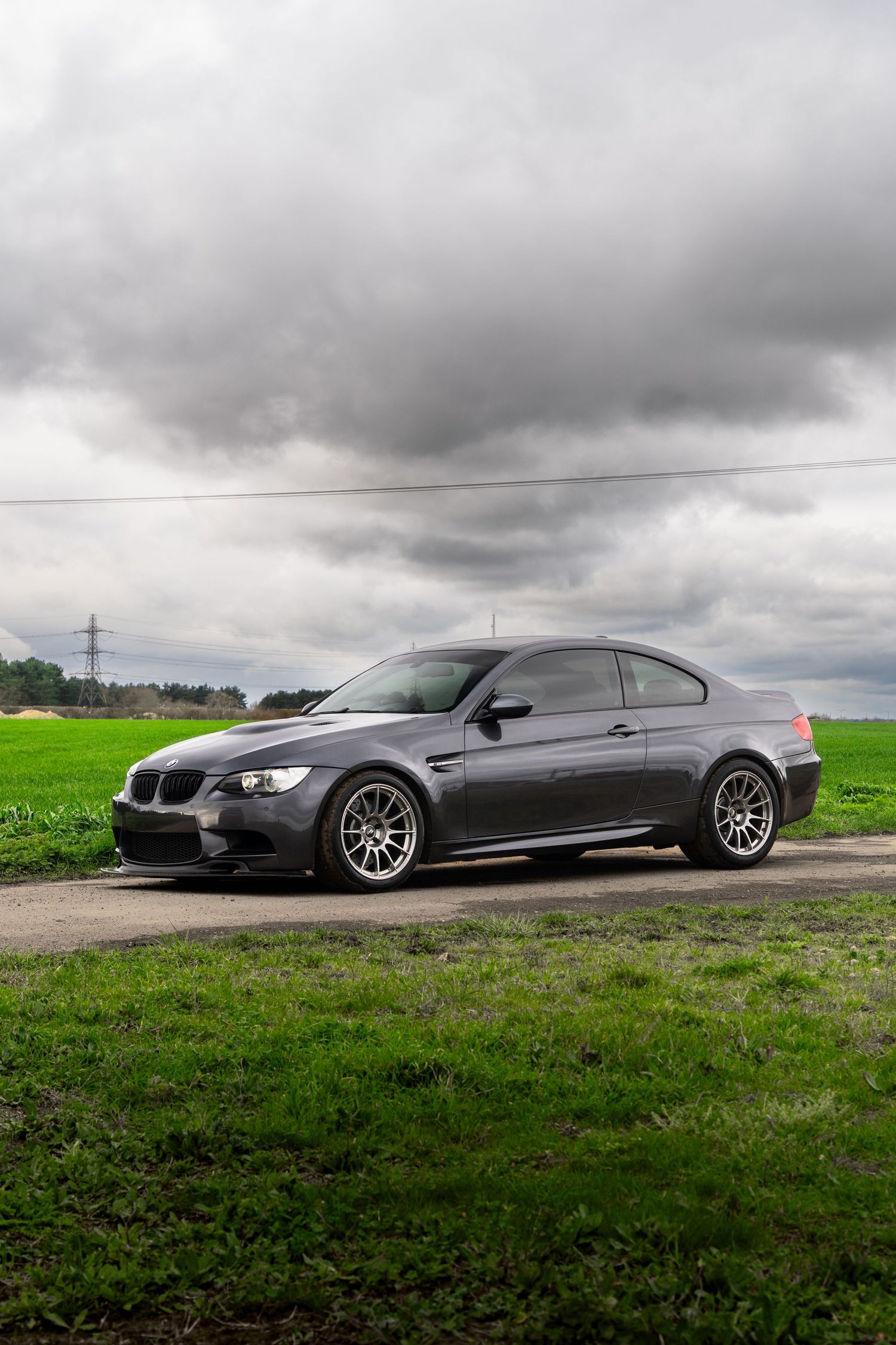 Dark gray BMW M3 coupe parked on a rural dirt path surrounded by green fields under dramatic cloudy skies.
