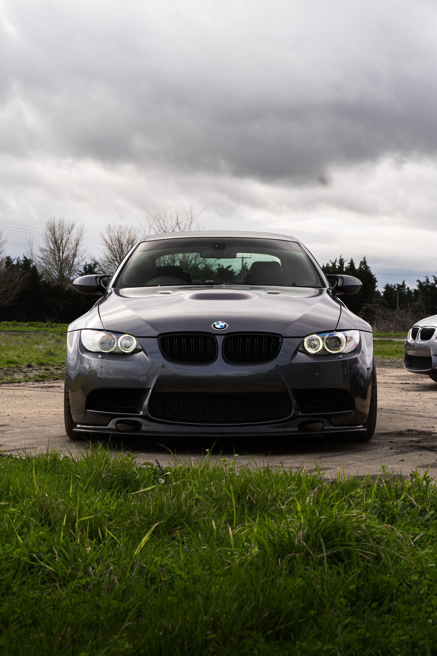 Front view of a modified dark gray BMW M3 E92 with angel eyes and aggressive front splitter on a cloudy day.