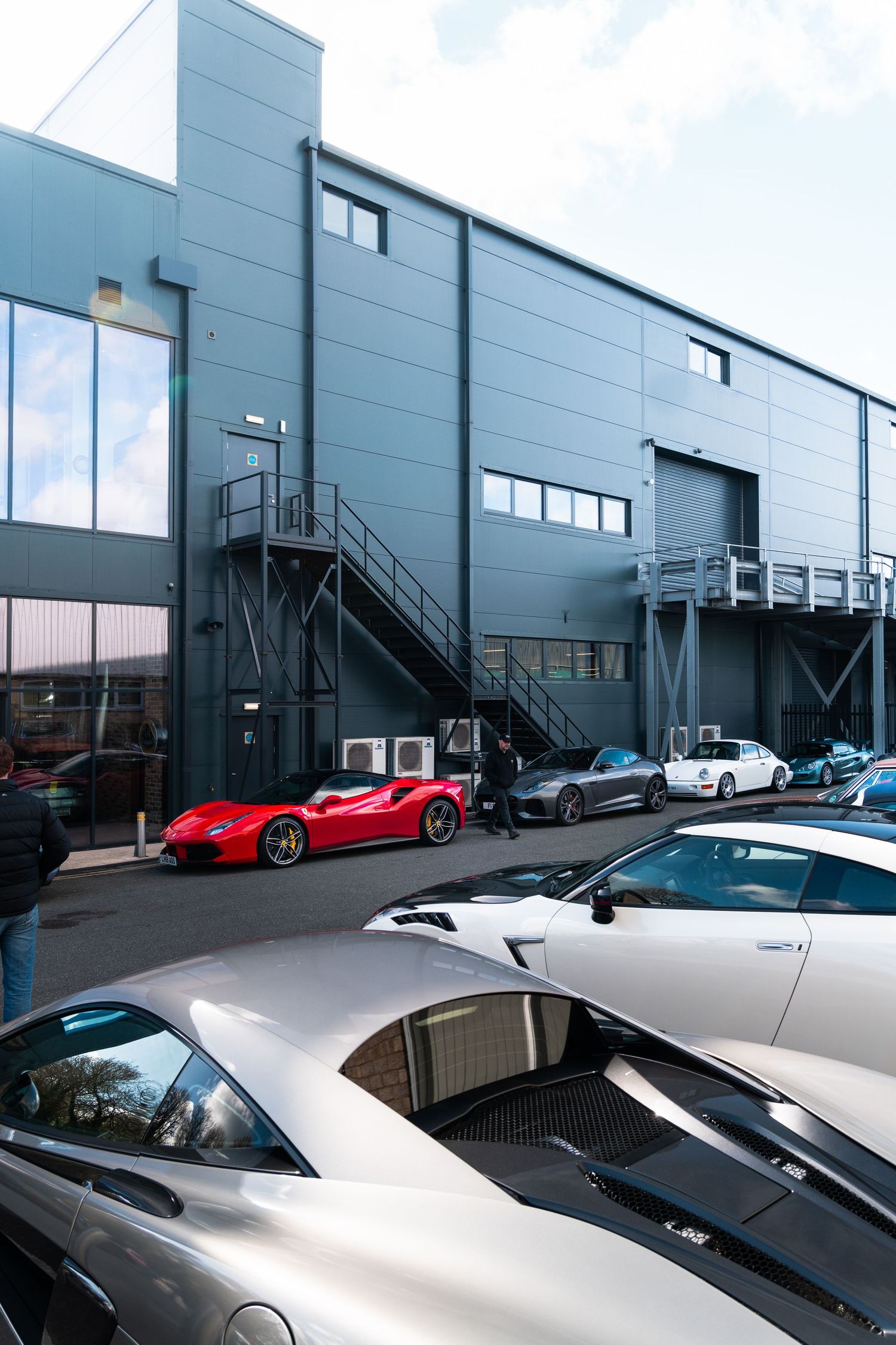 Collection of supercars including a red Ferrari parked outside a grey industrial building.