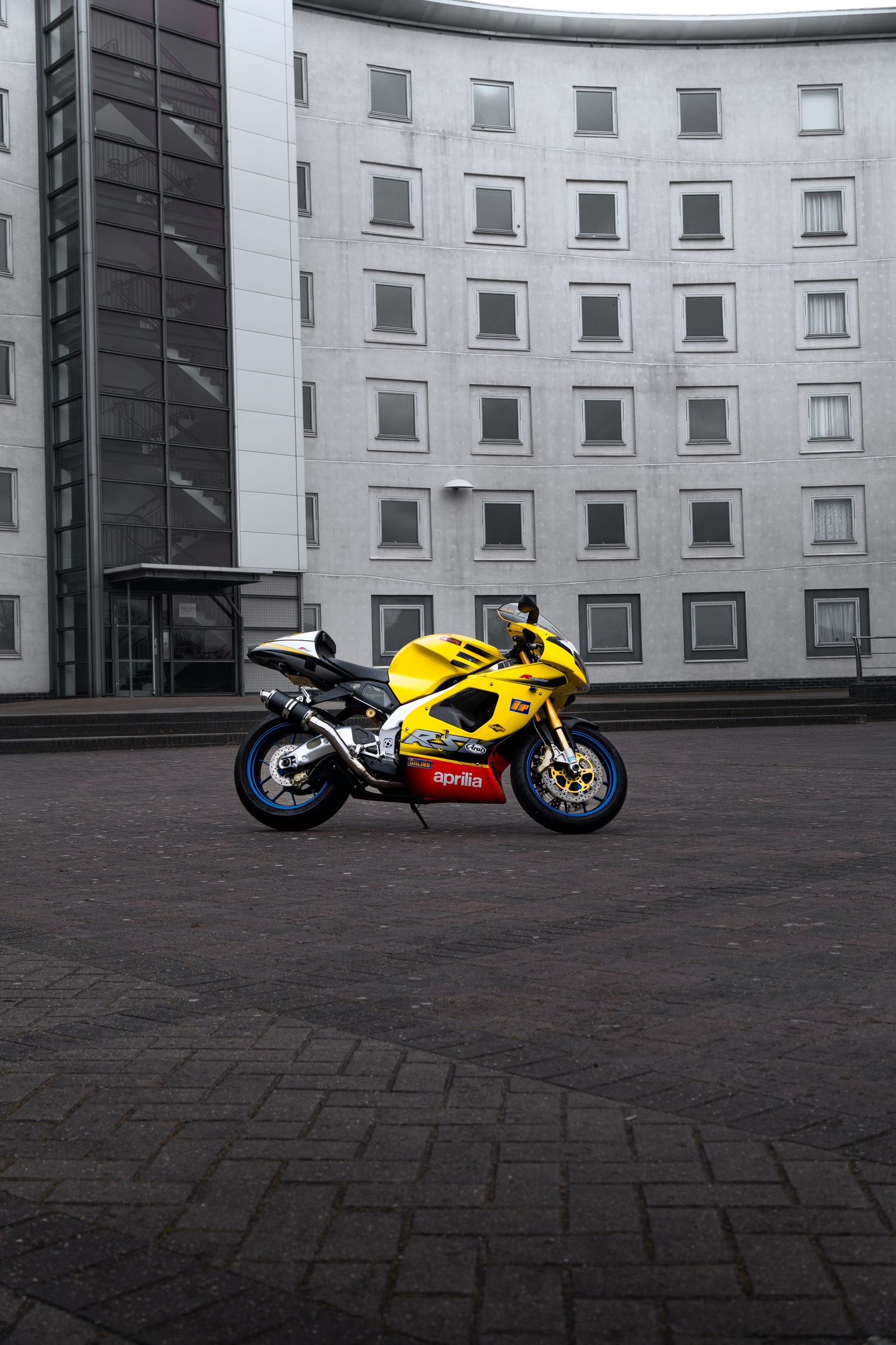 Yellow Aprilia RSV-R motorcycle parked on cobblestone plaza beside a curved multi-story building facade.
