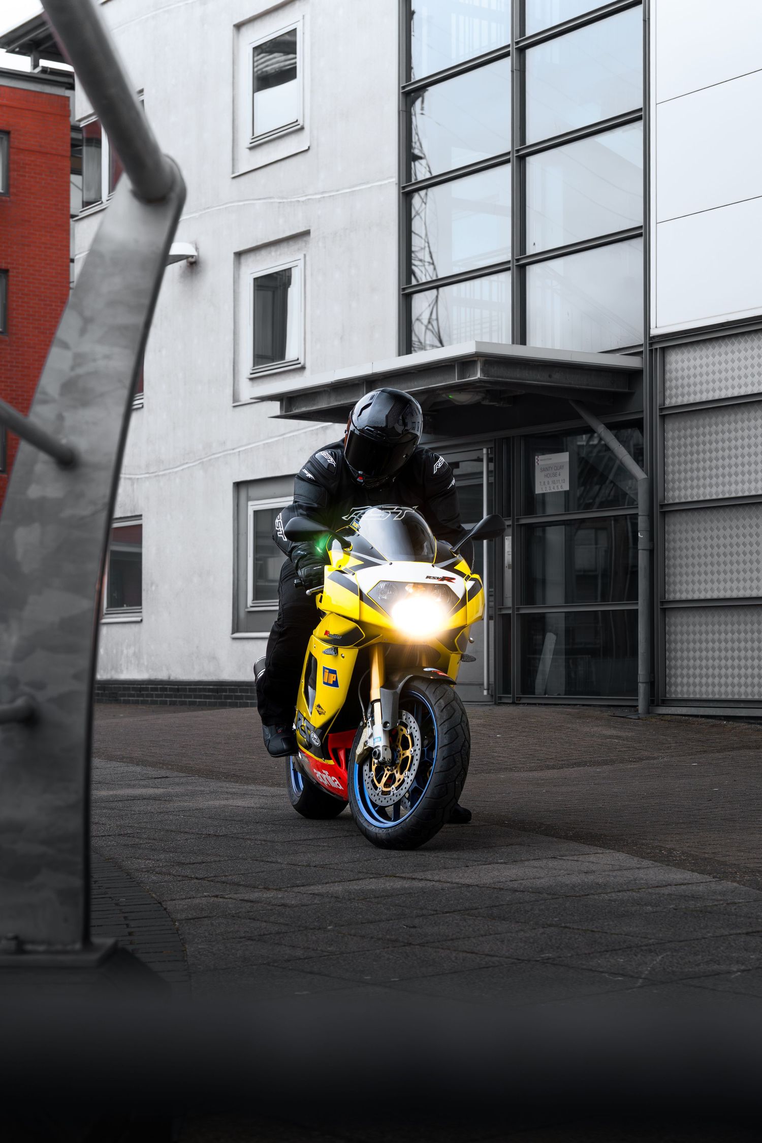 Yellow Aprilia sportbike with headlight on approaching building entrance at dusk.