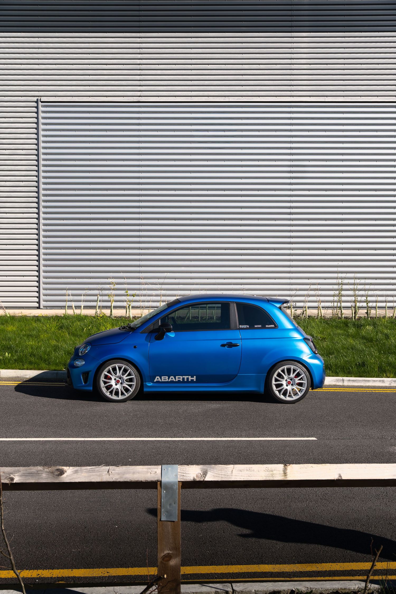 Blue Abarth 500 side profile shot against corrugated metal building wall with white multi-spoke alloy wheels.