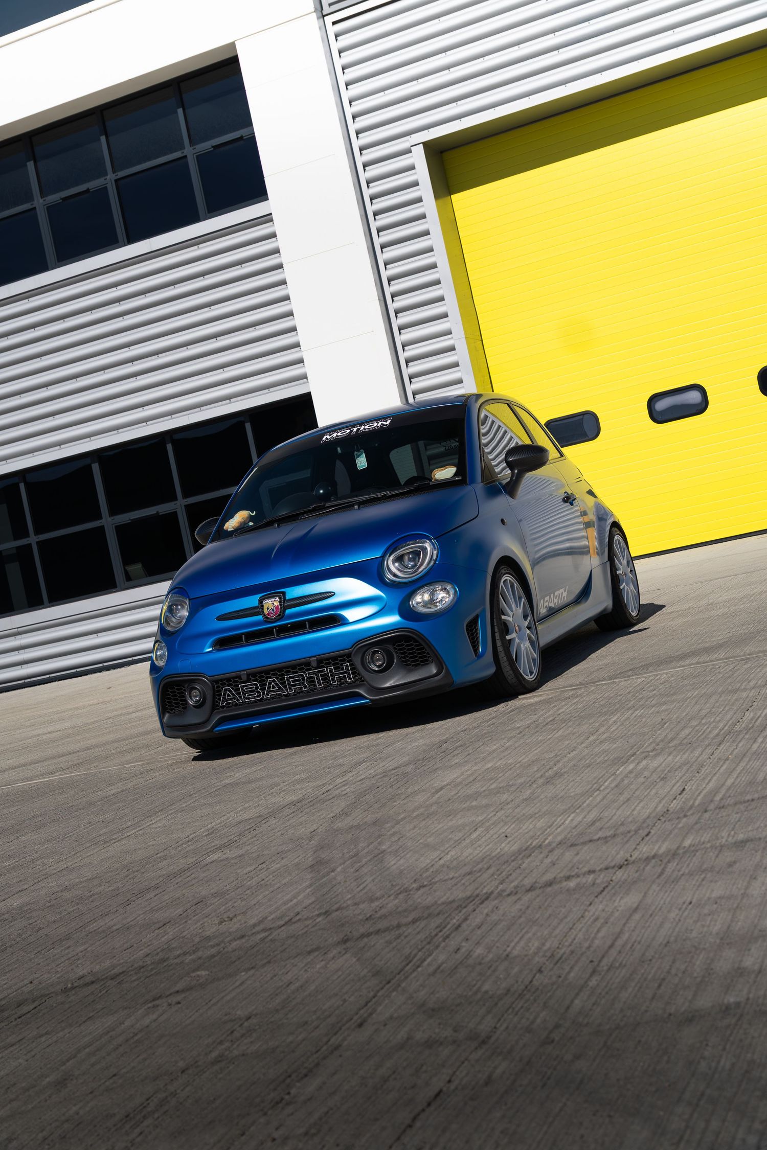 Blue Abarth 595 performance hatchback cornering dynamically by yellow garage doors.