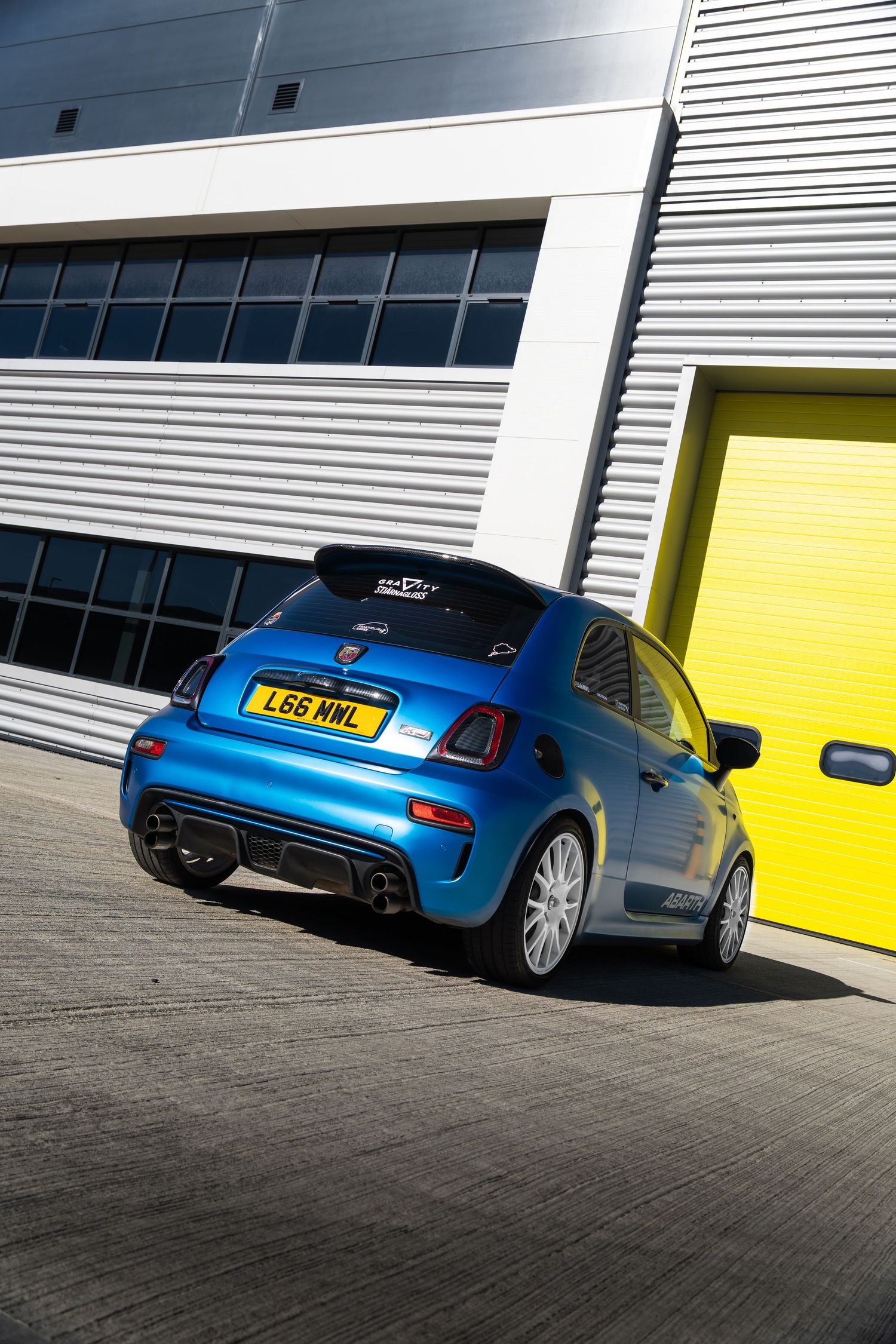 Blue Abarth 595 with dual exhausts and UK number plate outside warehouse with yellow door.