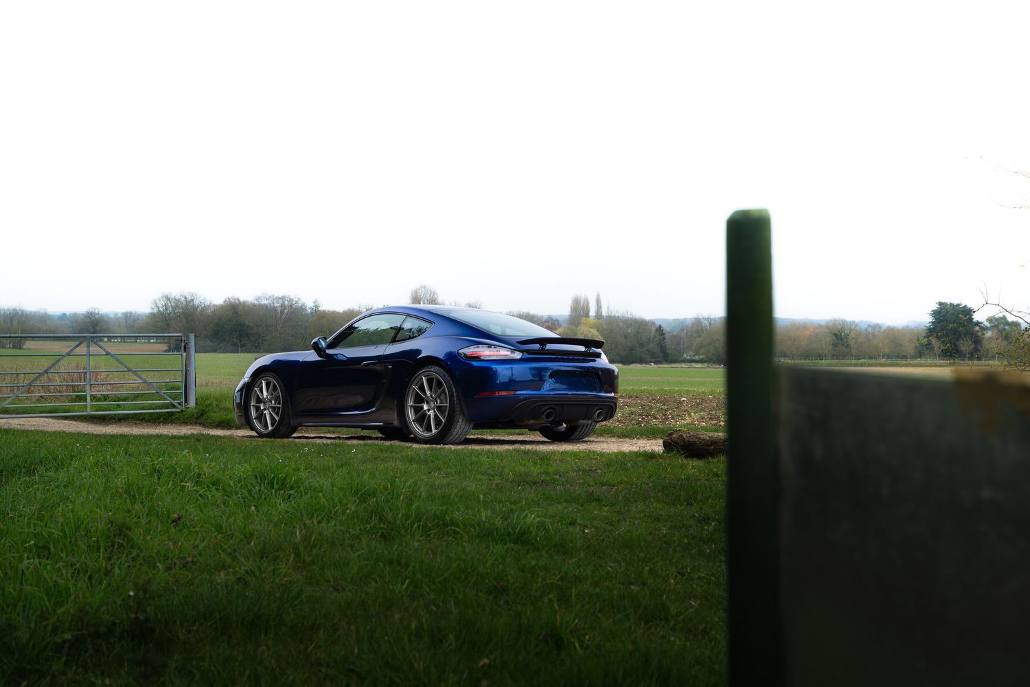 Blue Porsche 718 Cayman parked near a countryside gate with open green fields and pale sky.