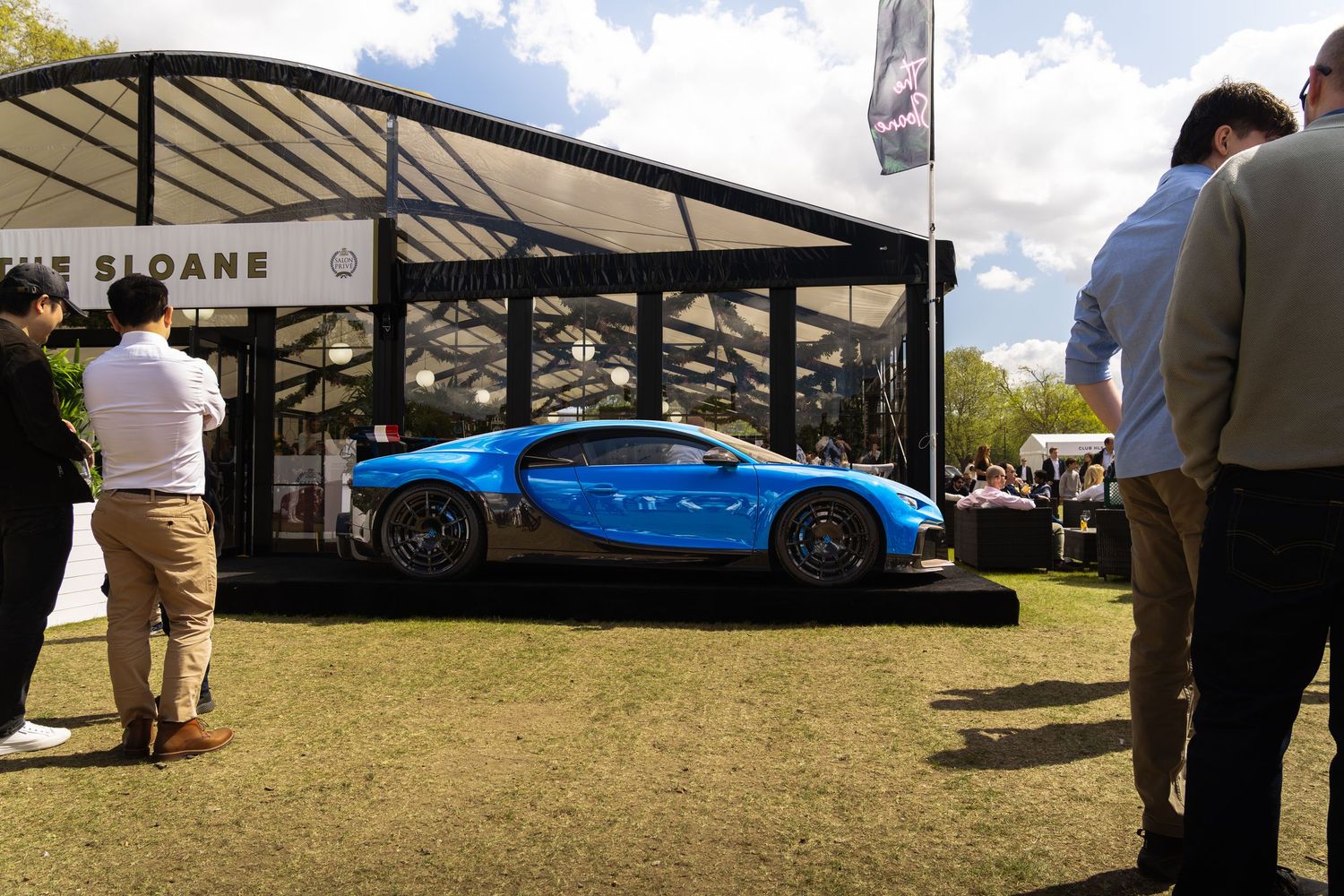 Sky blue Bugatti Chiron on elevated display outside Sloane dealership pavilion at luxury outdoor car event.