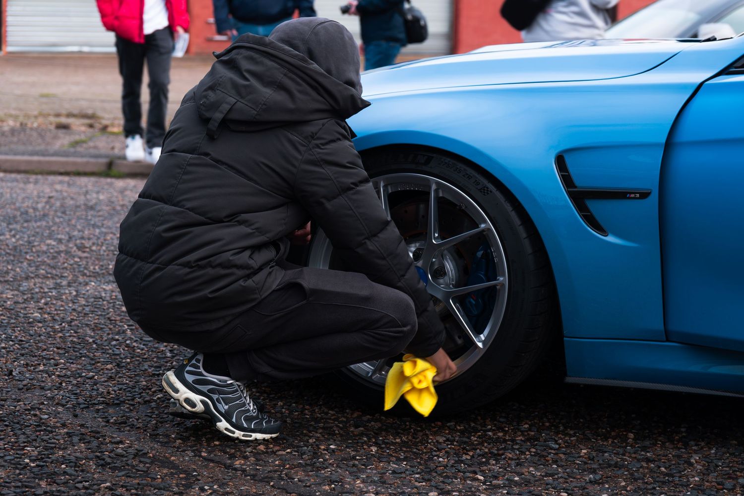 A person in black jacket crouches to clean the alloy wheel of a blue sports car.