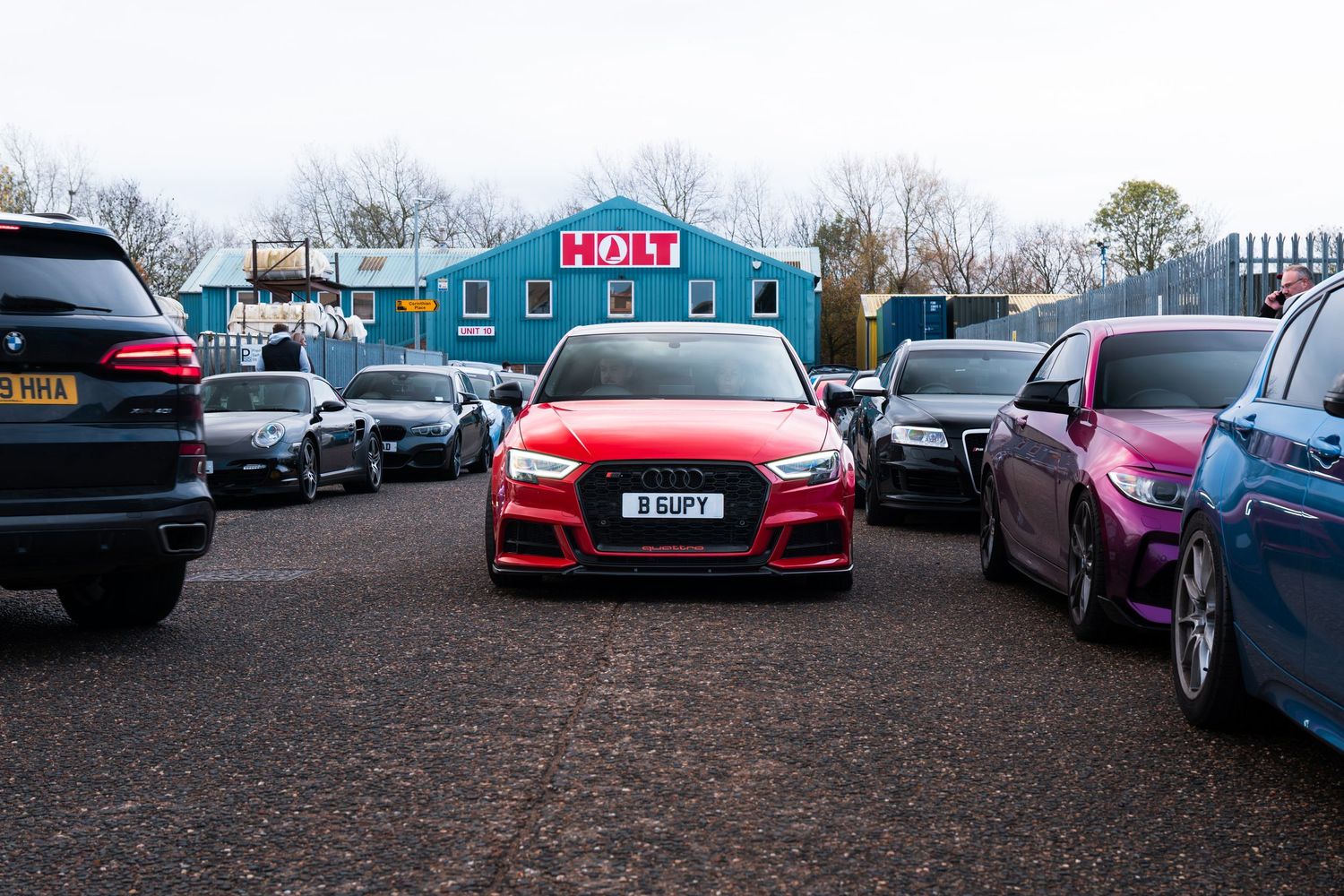 A red Audi RS3 parked at a busy outdoor car meet with colorful vehicles nearby.