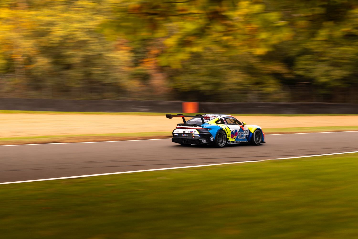 Colorful blue and yellow Porsche GT3 race car cornering on track with blurred autumn trees.