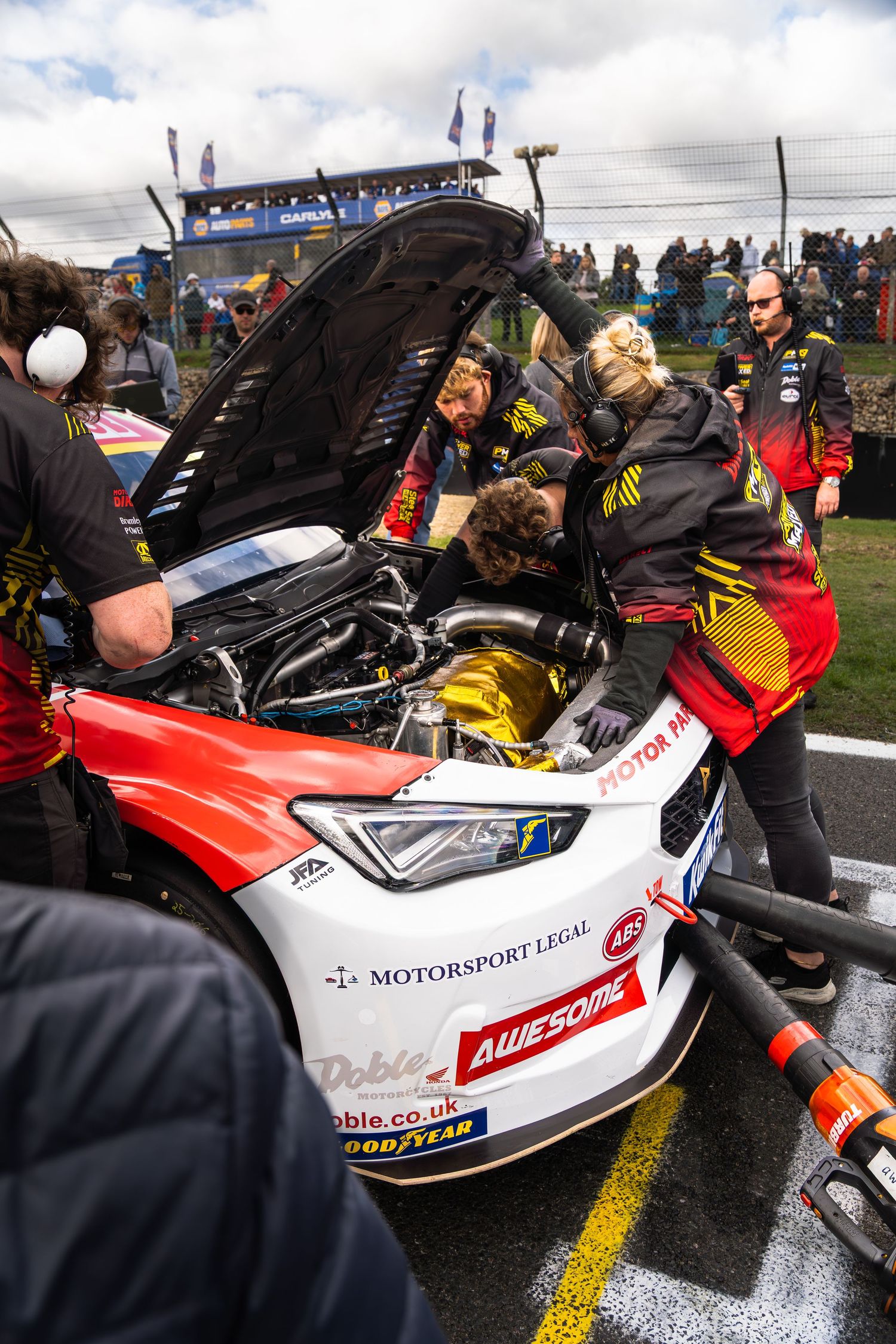 BTCC pit crew working on red and black race car engine with hood open during race event.