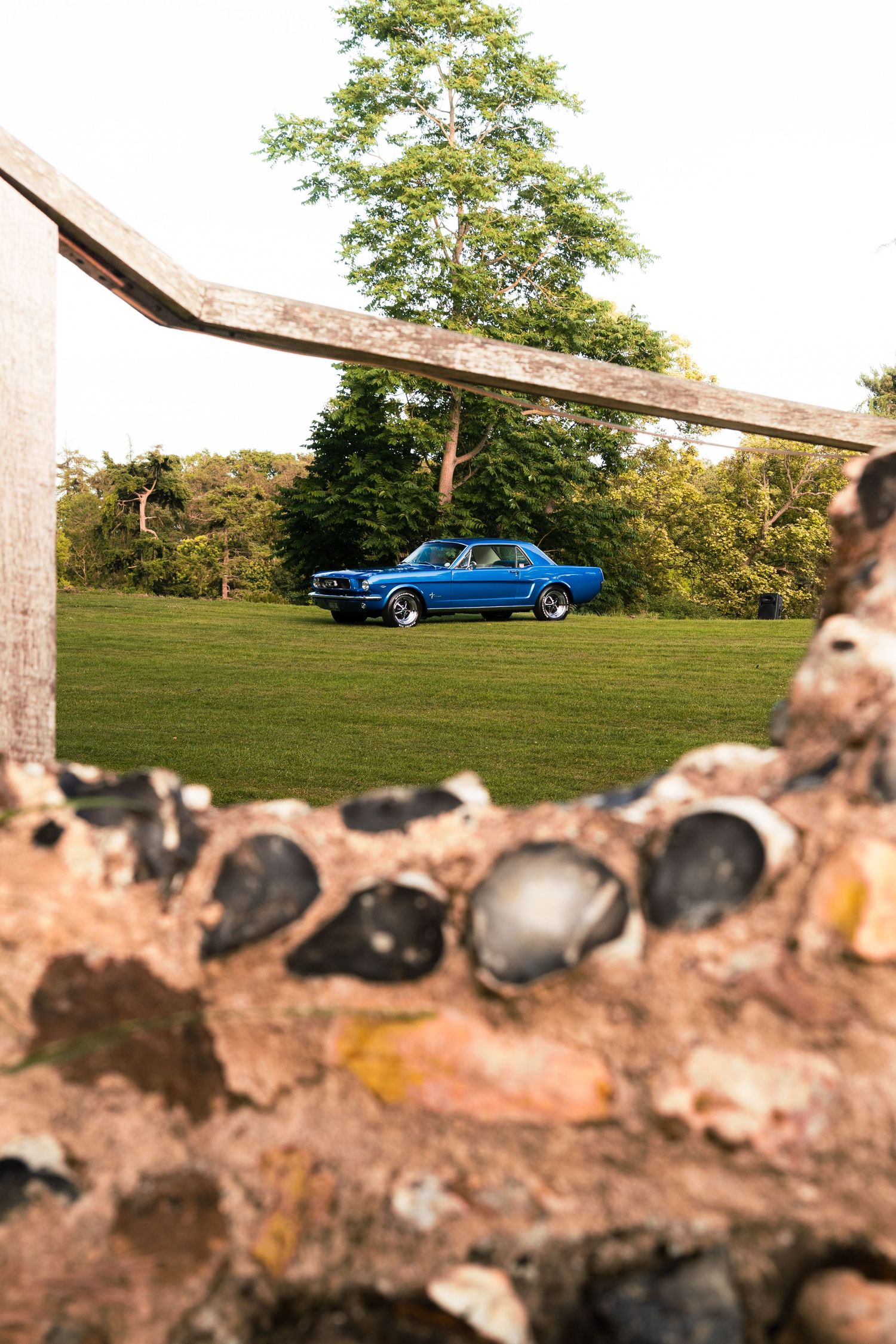 A classic blue Ford Mustang parked on green grass, framed by a rustic stone wall and wooden beam.