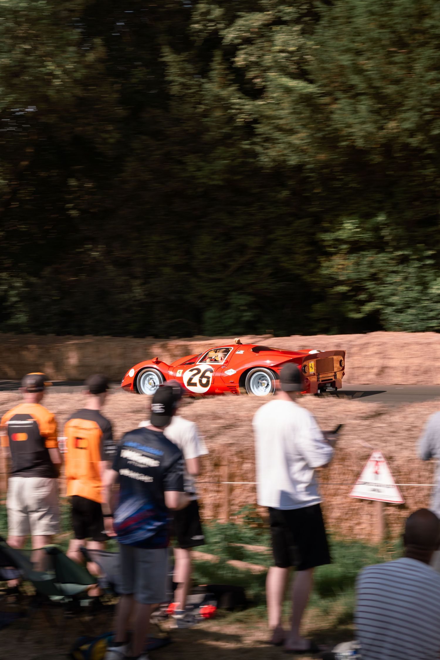 Red vintage race car number 26 speeds past hay bales as spectators watch at a classic motorsport hillclimb event.