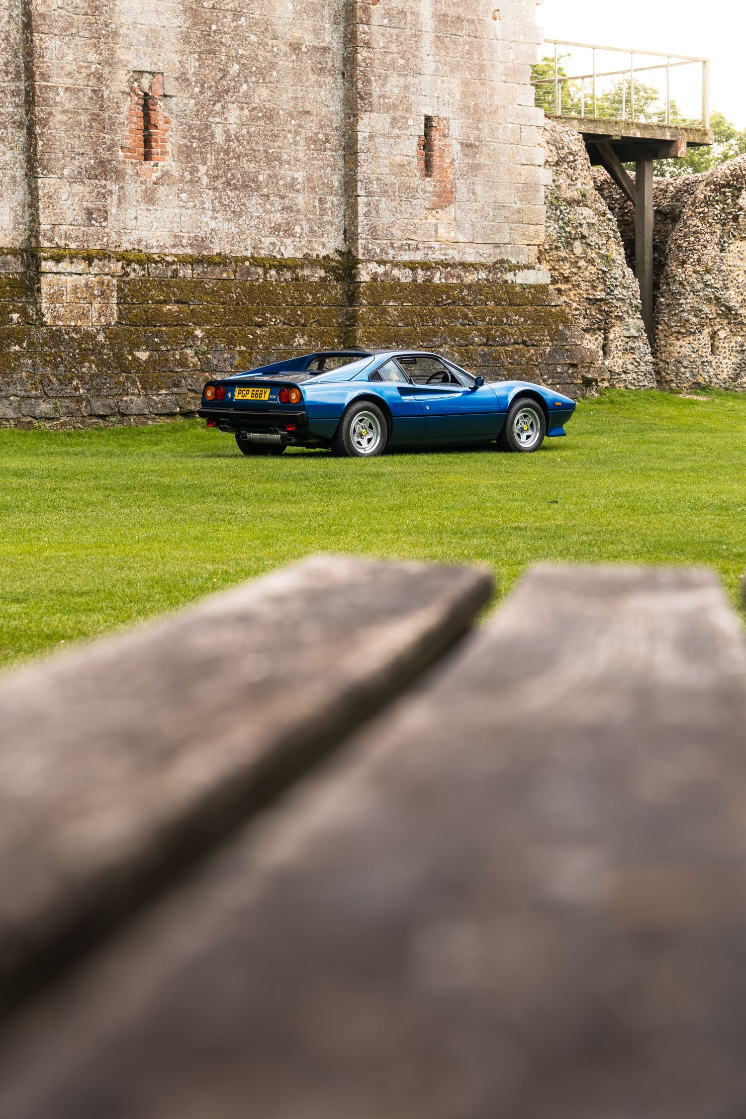 A classic blue Ferrari 308 parked on green grass beside ancient stone castle walls, viewed from behind a wooden bench.
