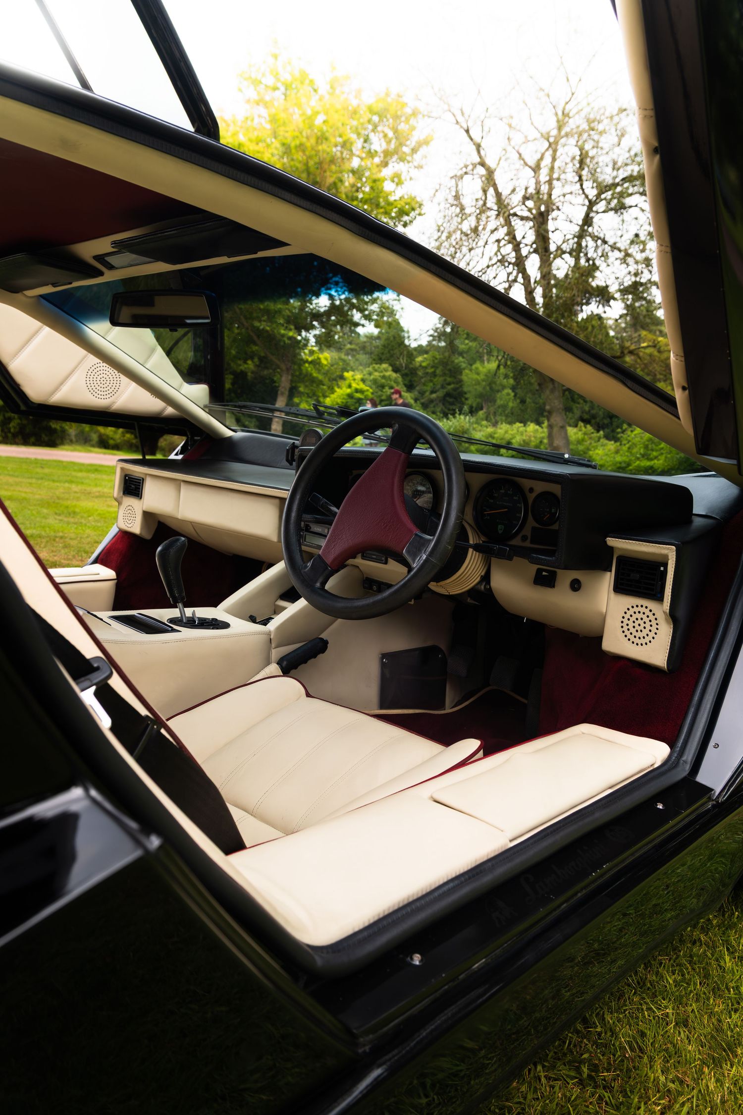 Vintage car interior with cream and red upholstery, steering wheel, and dashboard, door open revealing lush green garden.