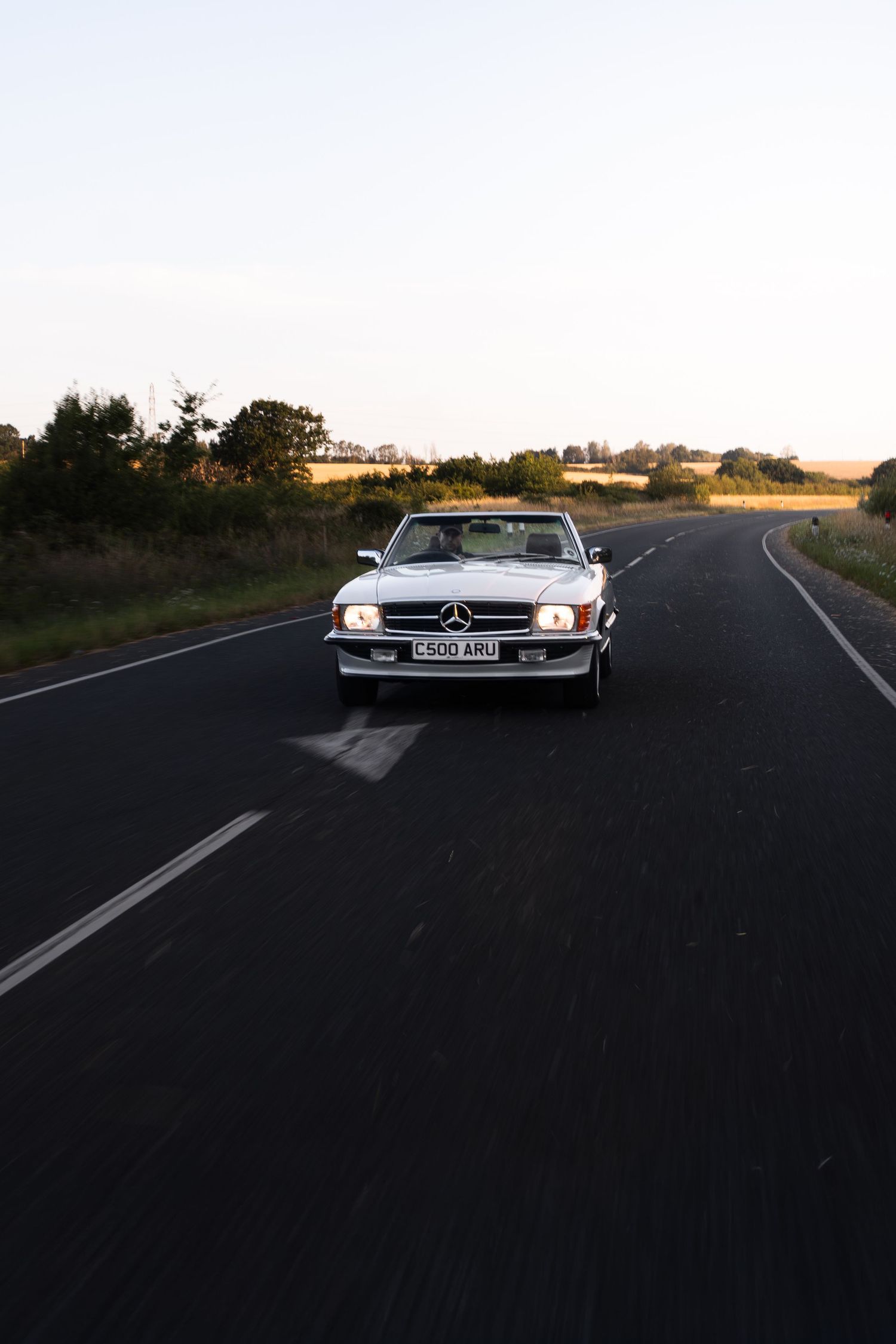 Classic white Mercedes-Benz convertible driving on a rural road at dusk with headlights on.