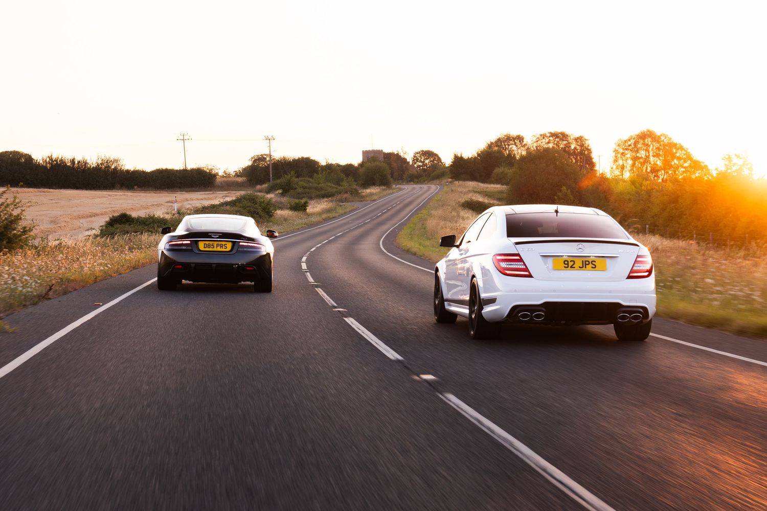 Two luxury cars driving on a winding country road at golden sunset, white Mercedes-Benz and dark sports car.