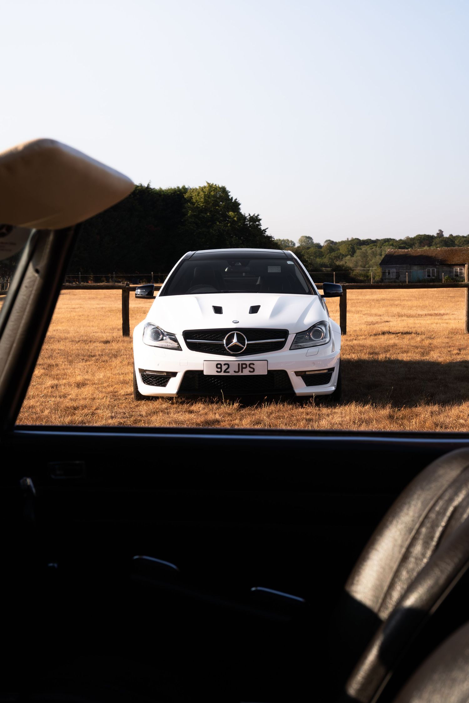 White Mercedes-Benz AMG coupe with plate 92JP5 parked in a golden field, viewed through an open car door.